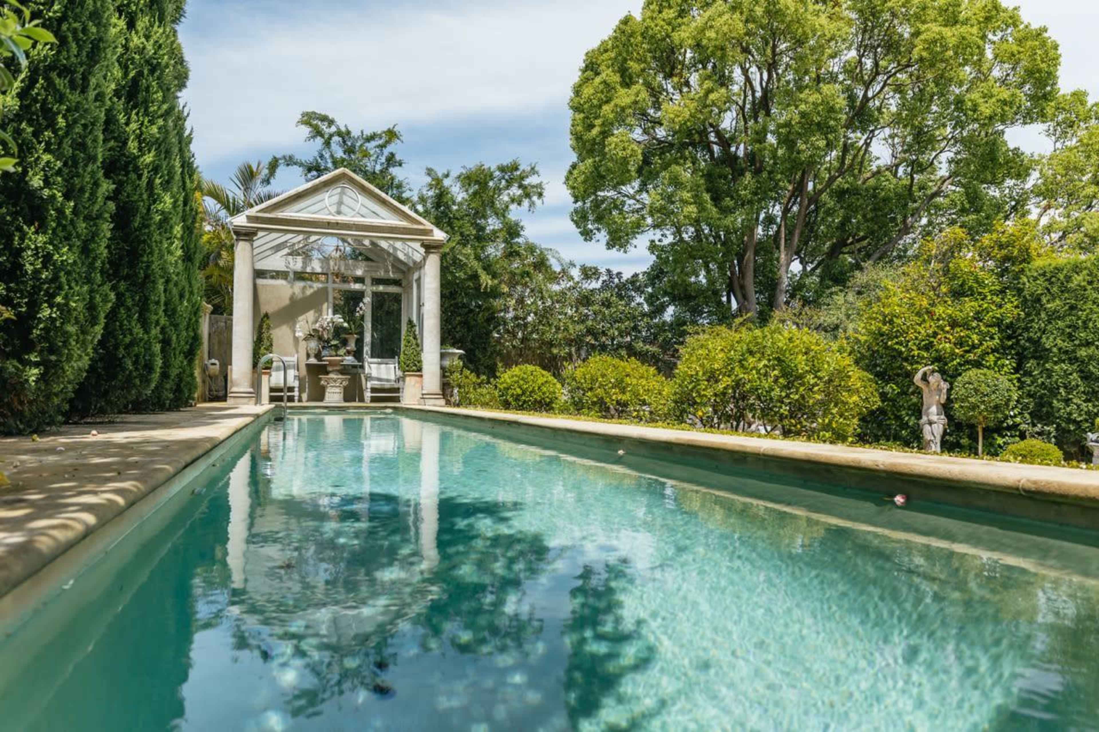 A clear swimming pool bordered by lush greenery and a glass conservatory in the background.