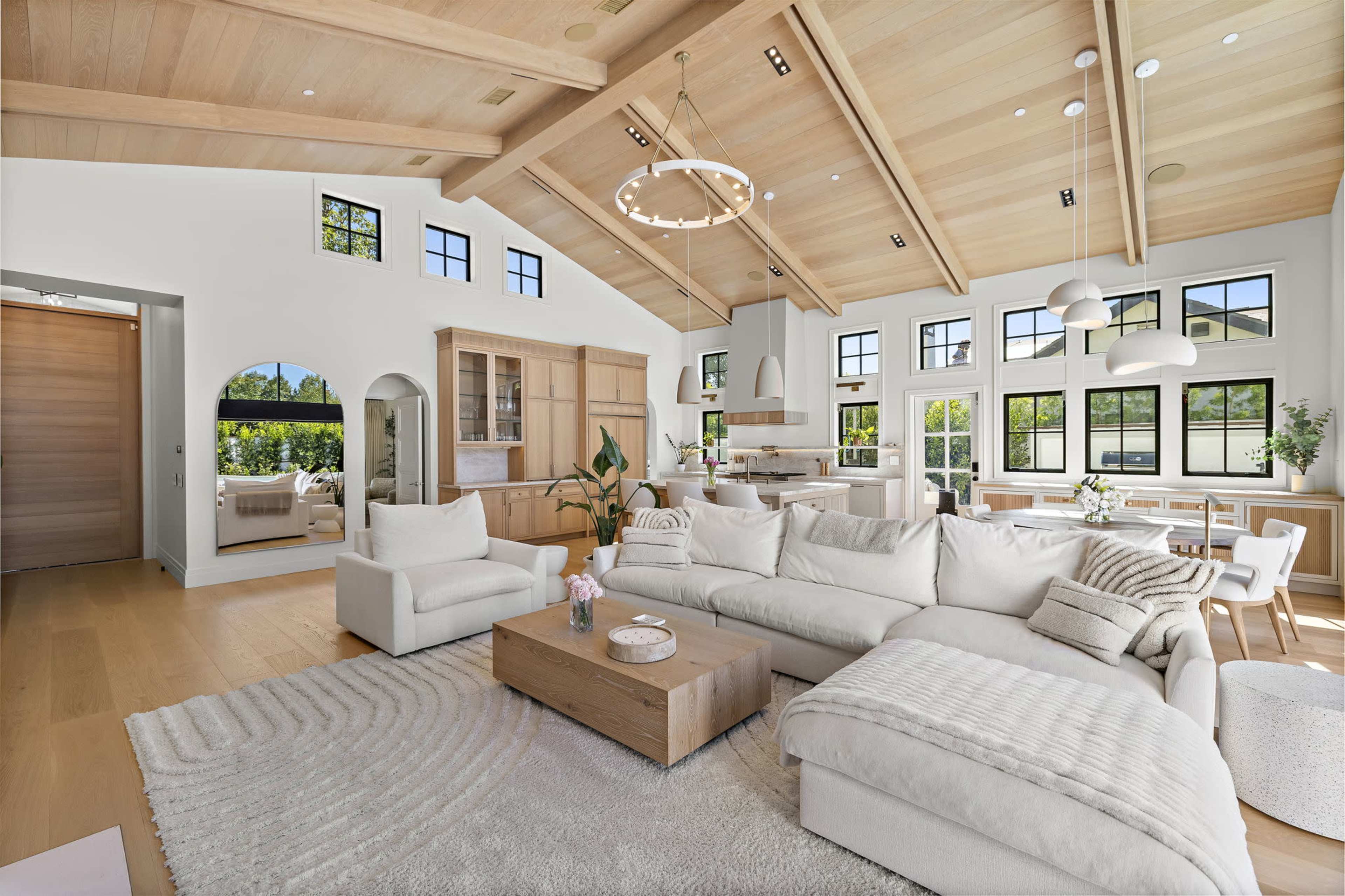 The image shows a spacious, modern living room with a high ceiling, large windows, and a light color palette featuring a white sectional sofa, a wooden coffee table, and a dining area in the background.