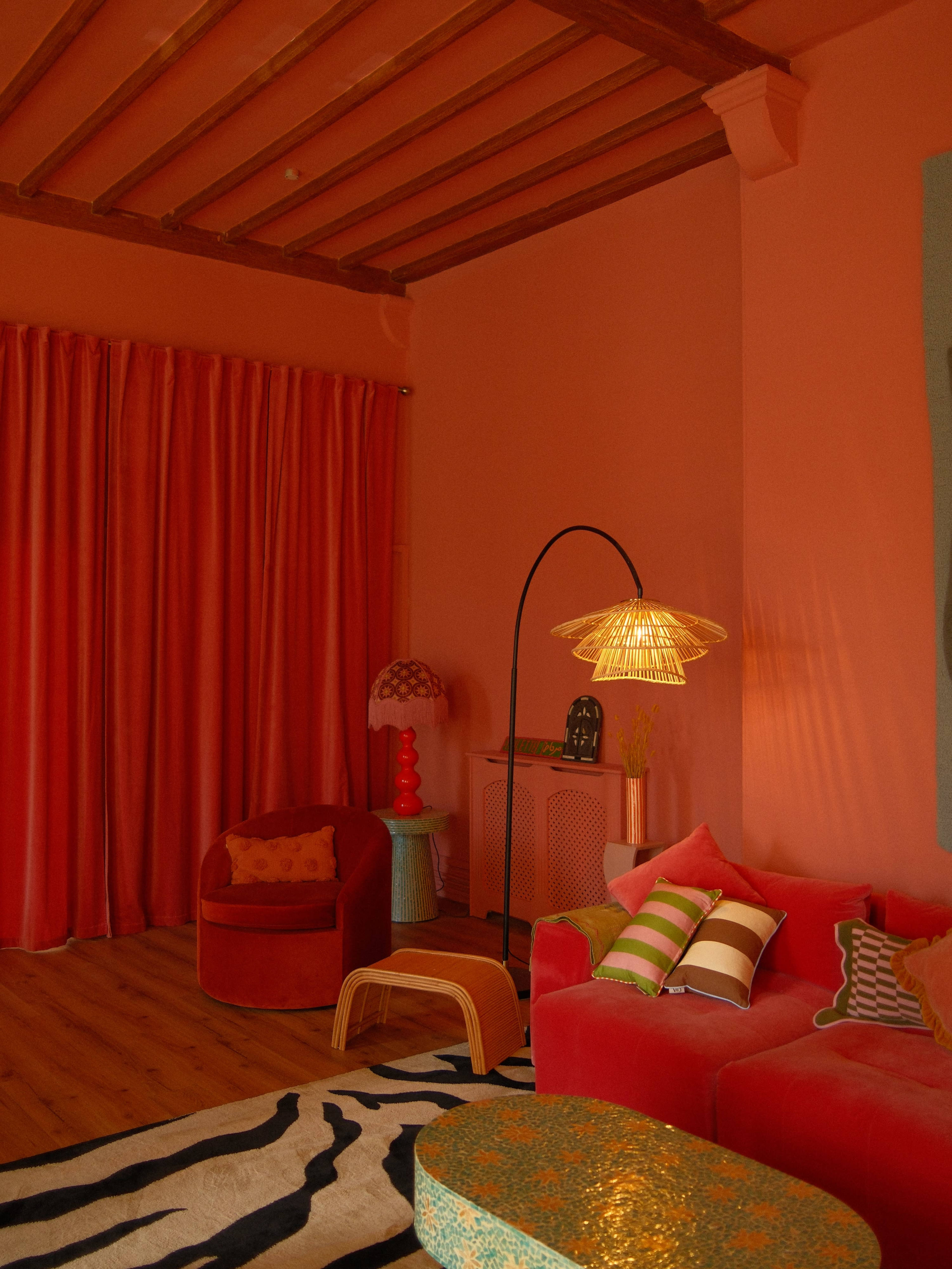 The scene shows a vibrant interior room with coral pink walls, a wooden ceiling, plush seating in various shades of red, and decorative elements like a floor lamp and curtains.