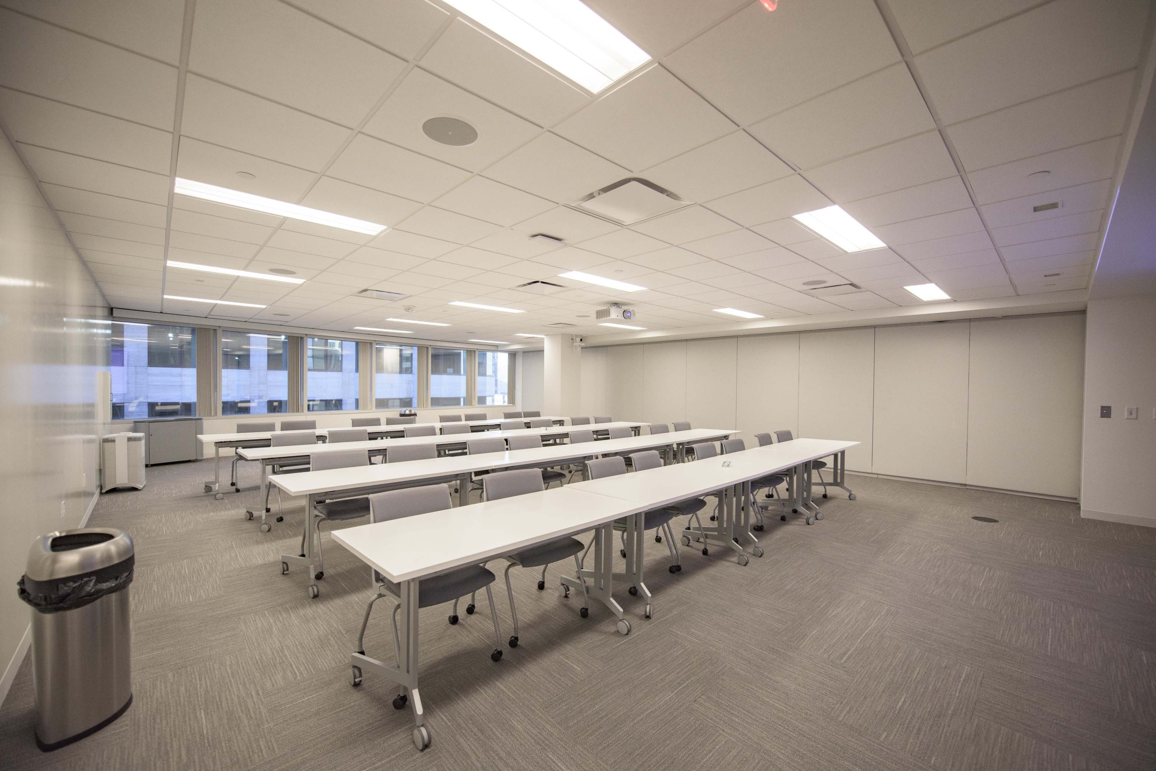 A conference room with multiple long tables arranged in a grid, equipped with chairs, and large windows allowing natural light.