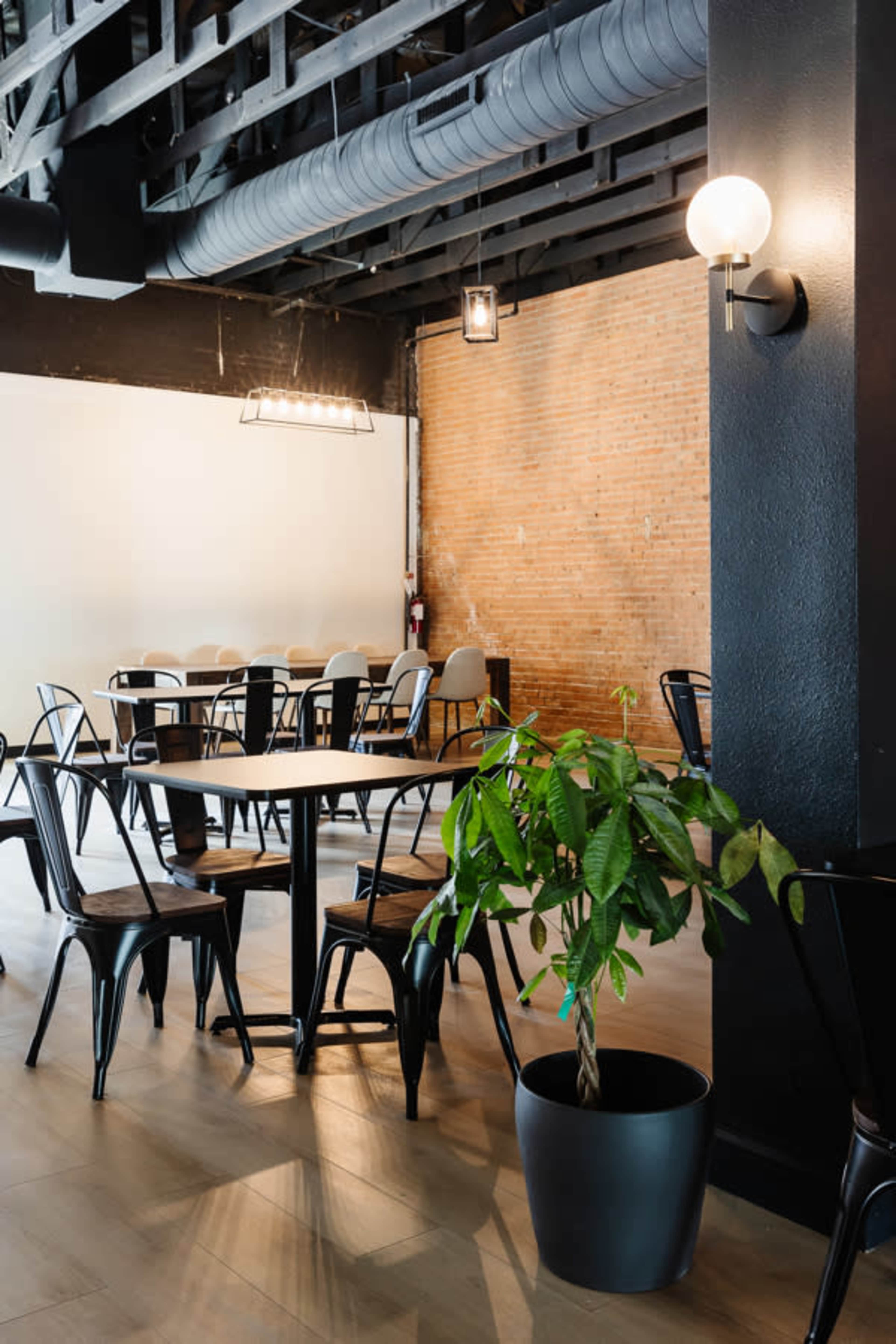 A modern dining area features black metal chairs and tables, a potted plant, exposed brick walls, and hanging light fixtures.