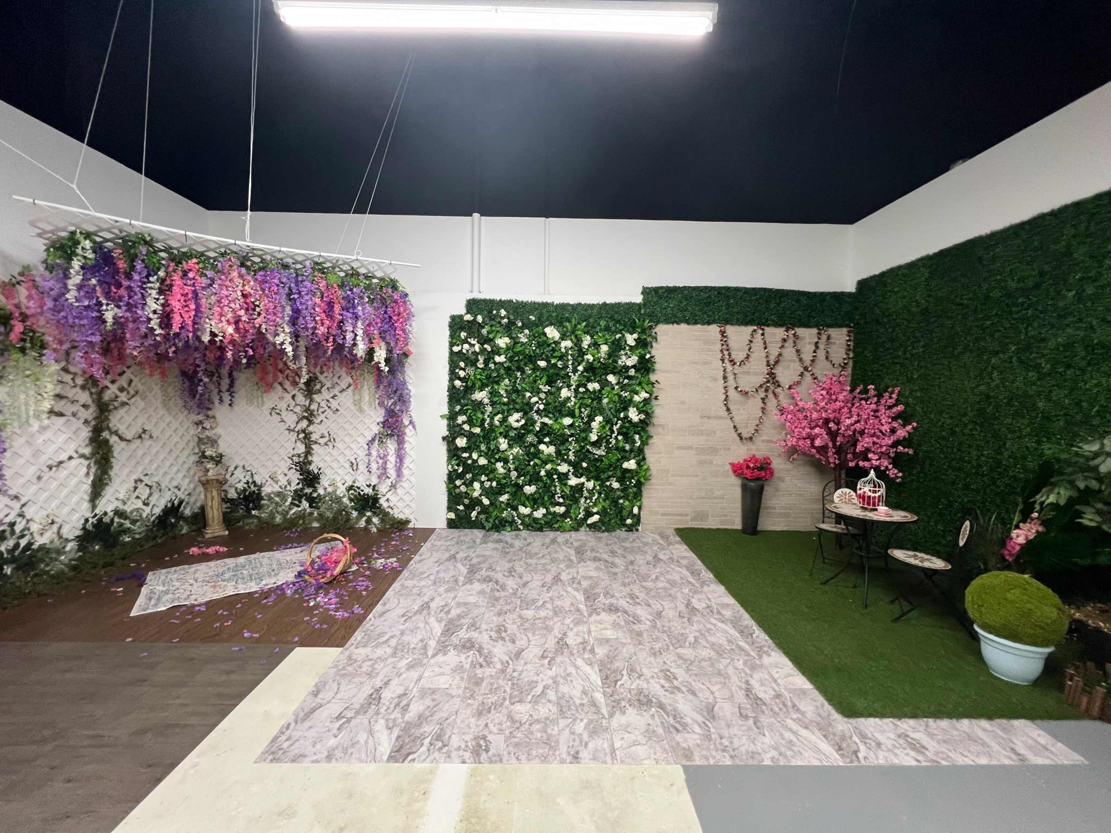 The image shows a staged indoor space with floral decorations, including hanging vines and a wall covered in white flowers, along with various textures like grass and stone tiles.
