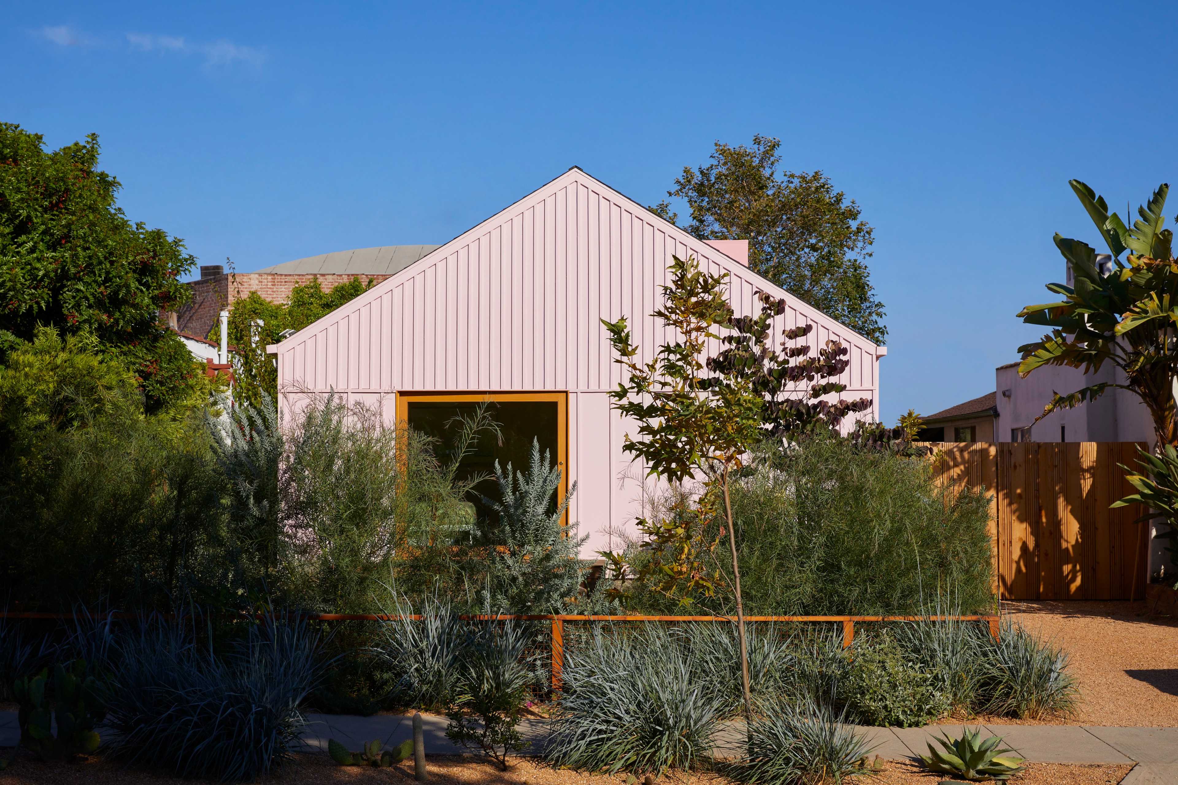 A pink-painted house with a peaked roof is surrounded by various green plants and a wooden fence.