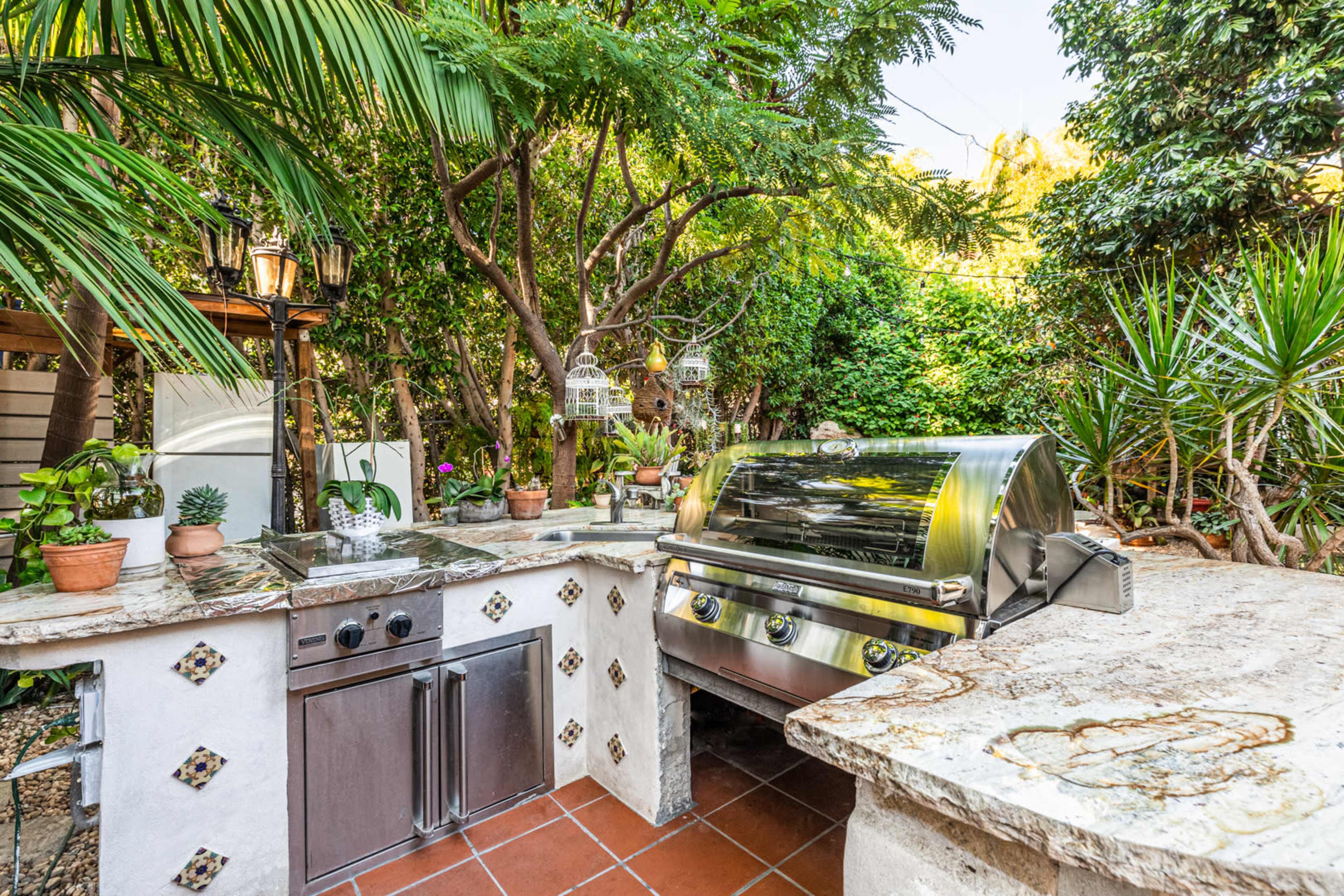 An outdoor kitchen featuring a stainless steel grill, stone countertops, and surrounded by lush greenery and potted plants.
