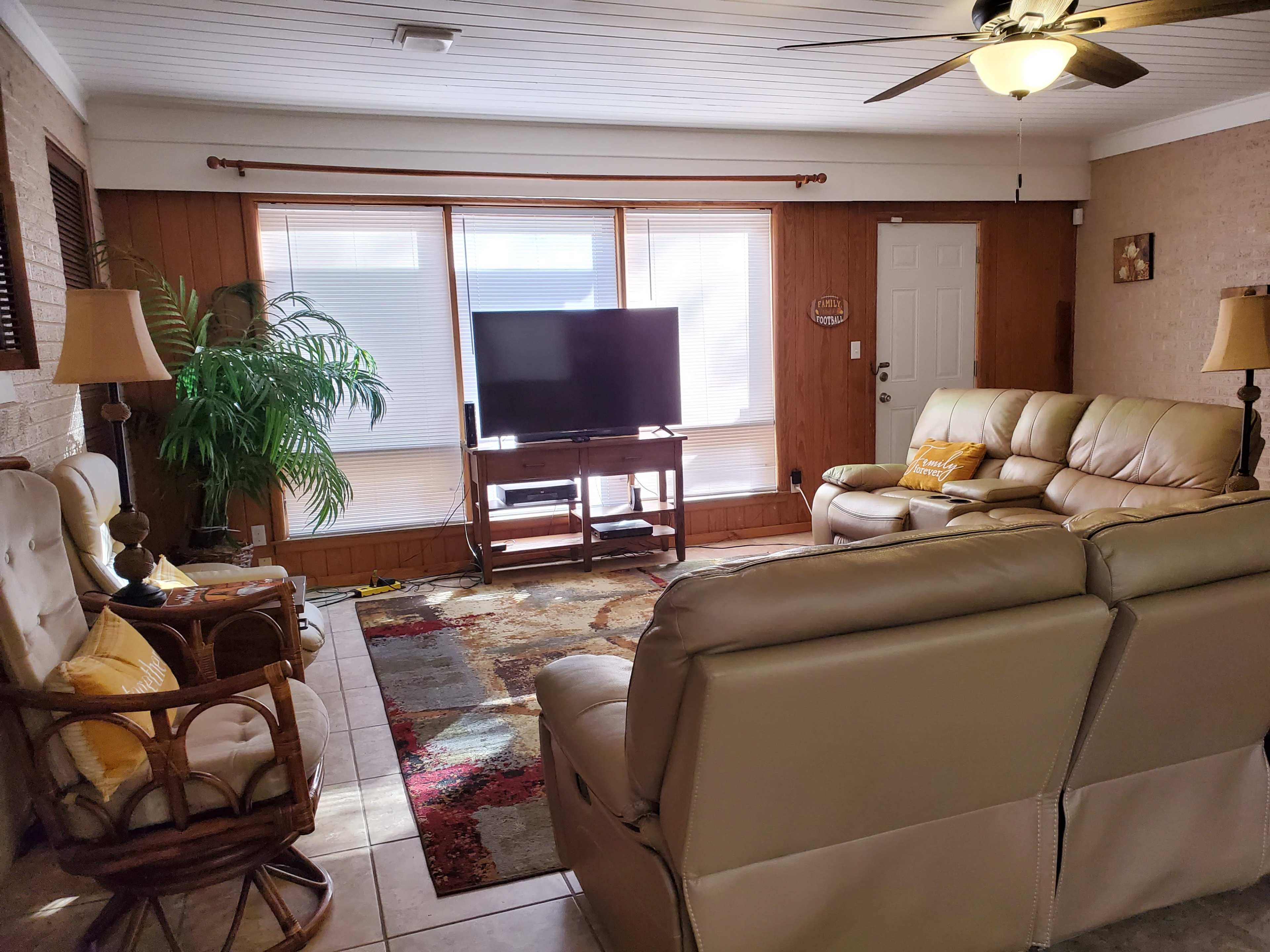 A living room features a television, beige reclining sofas, a rocking chair, and a large window with natural light.