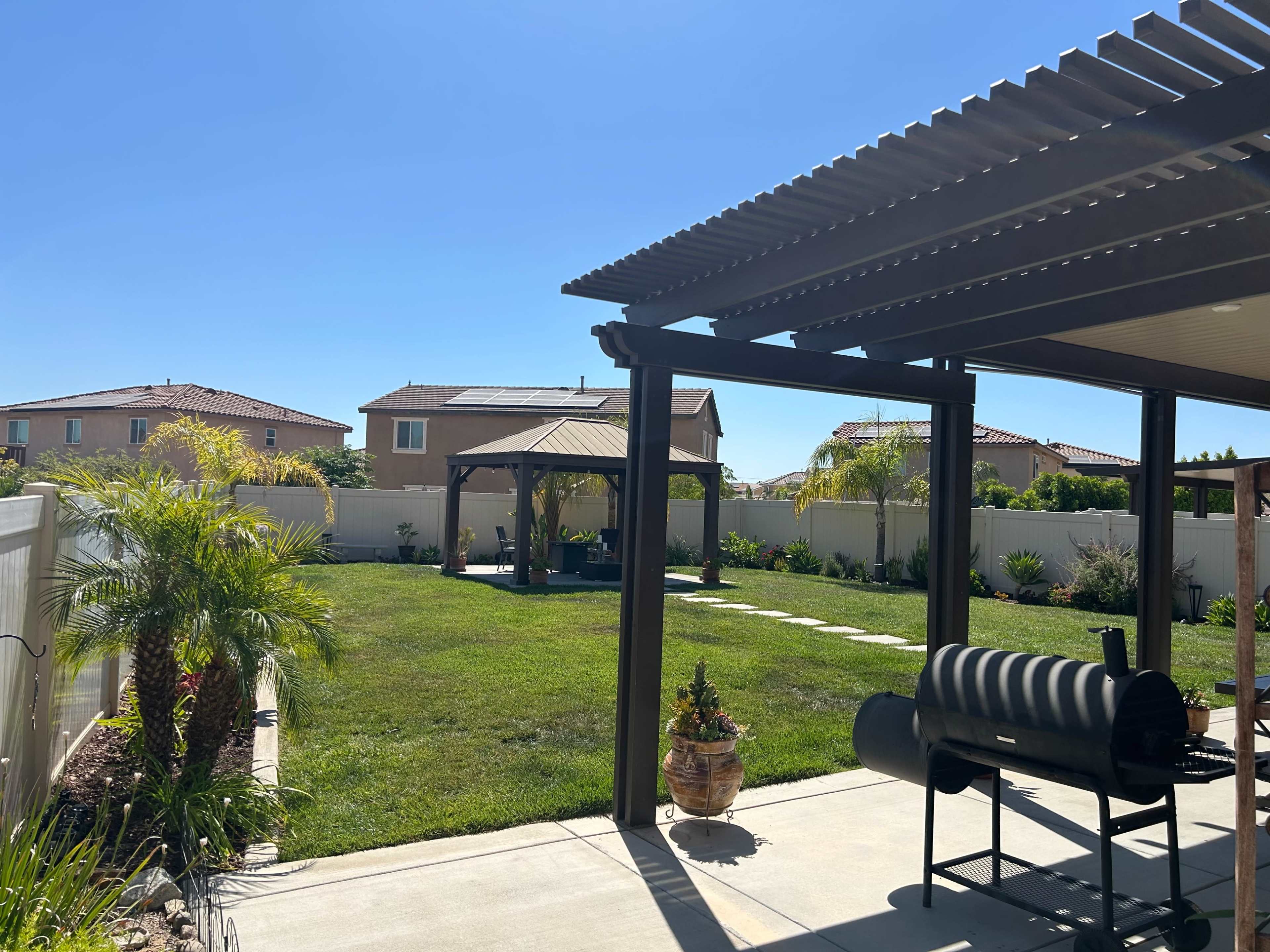 The image shows a landscaped backyard with a grass lawn, a gazebo in the distance, and a grill under a pergola.
