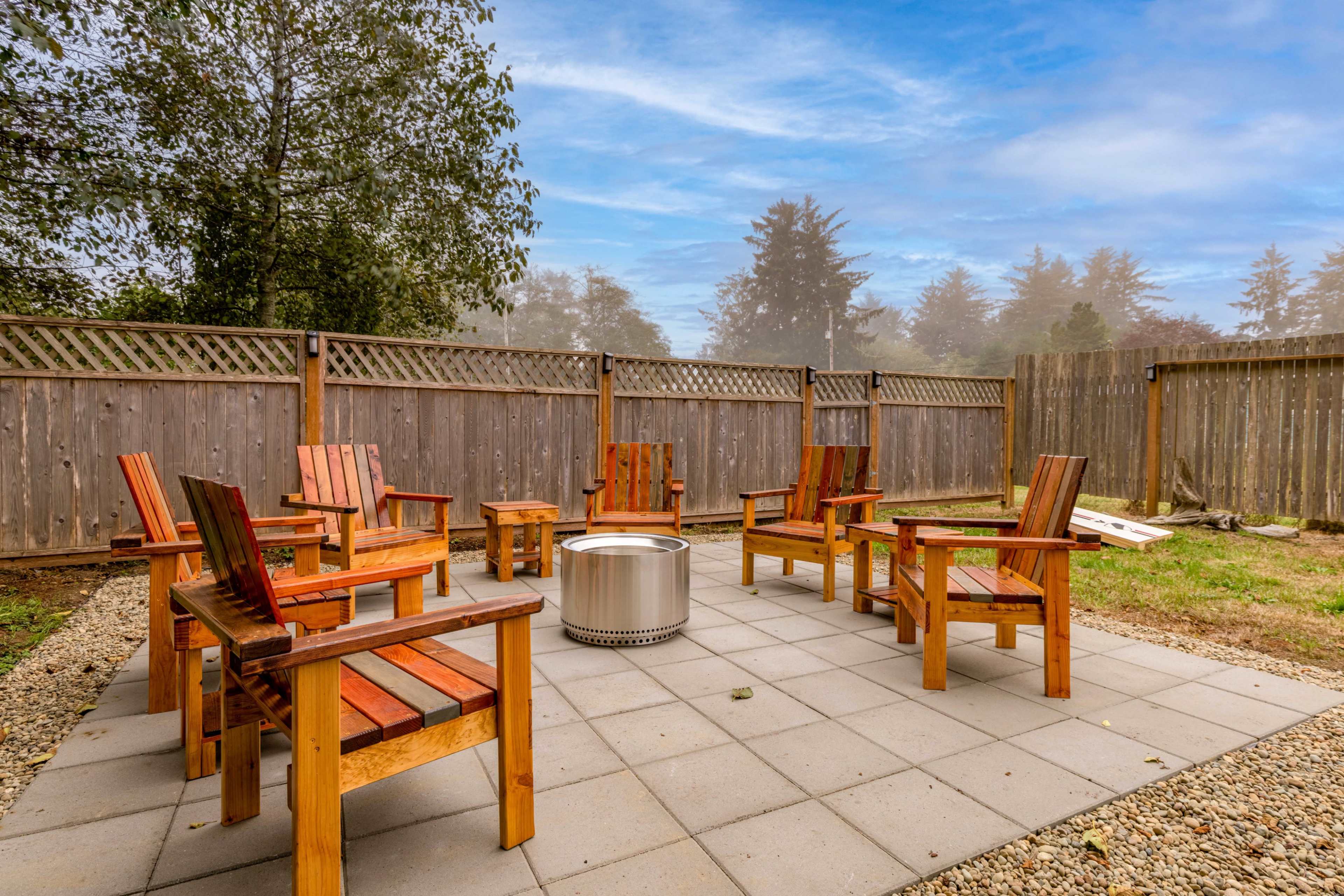 A circular seating area with wooden chairs surrounds a fire pit on a stone patio, enclosed by wooden fencing and trees in the background.