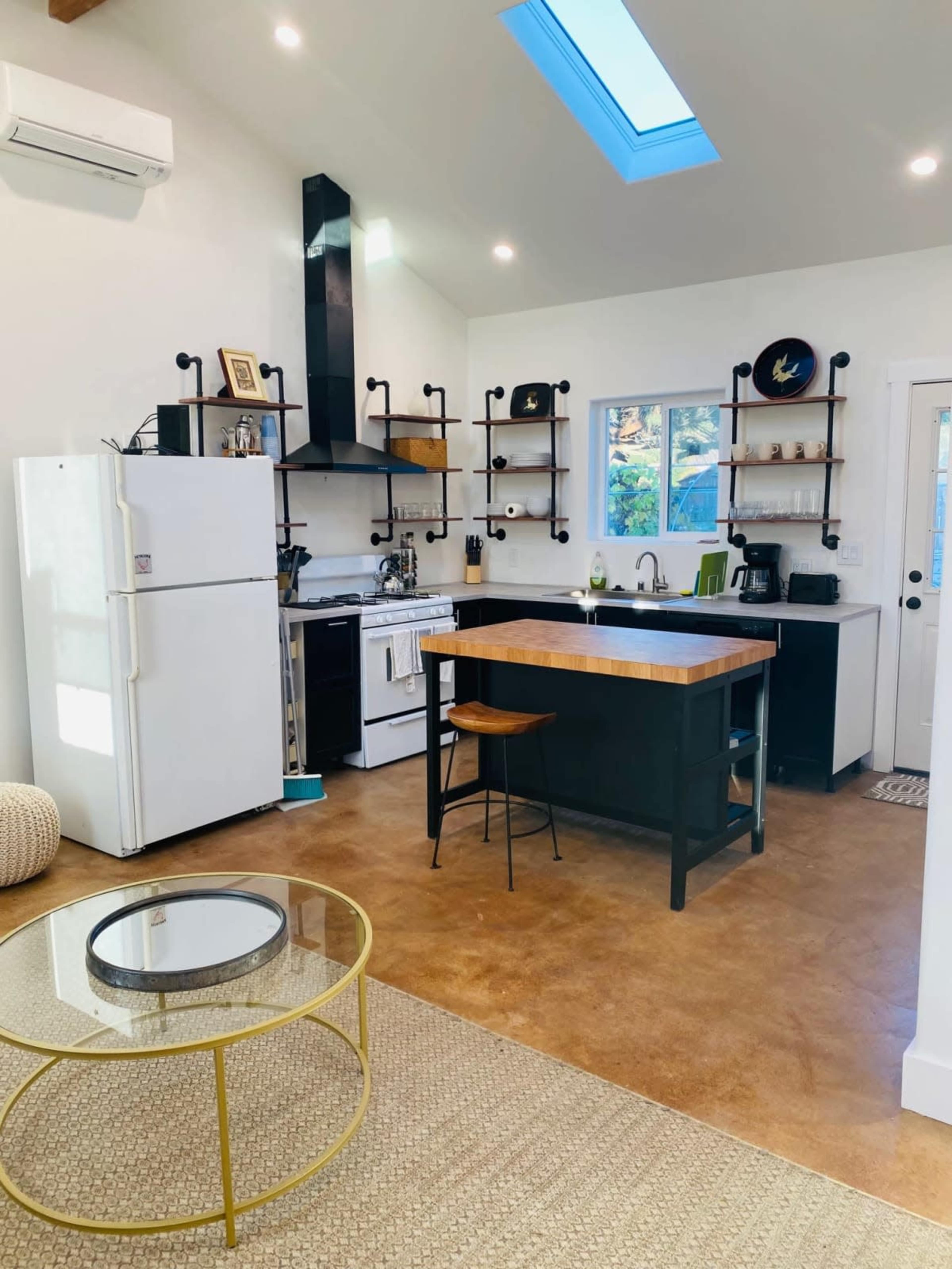 A modern kitchen features white cabinets, a black countertop, shelves with kitchenware, and a skylight above.