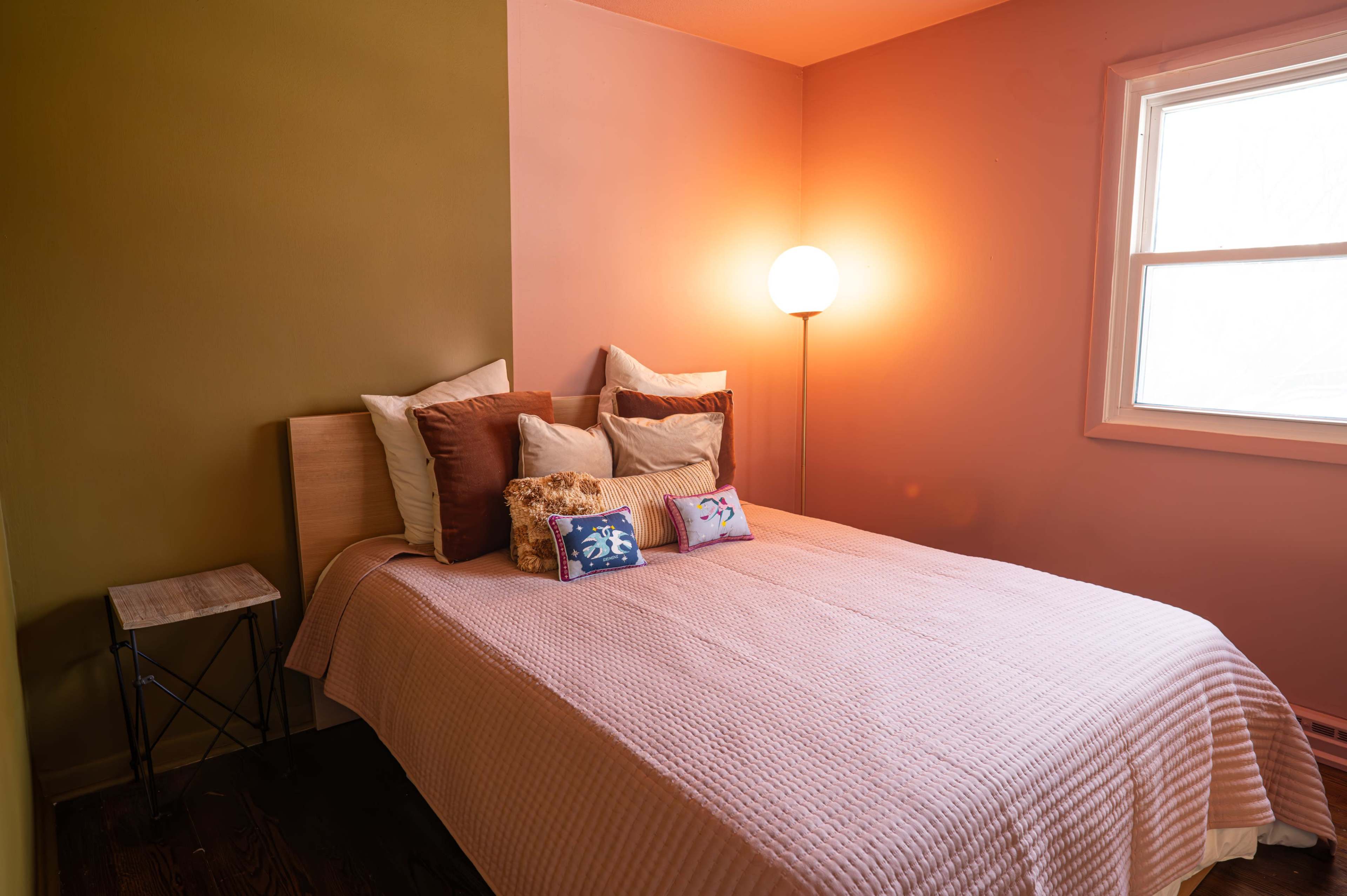 The image shows a bedroom featuring a bed with decorative pillows, a small side table, and a lamp next to a window with soft lighting.