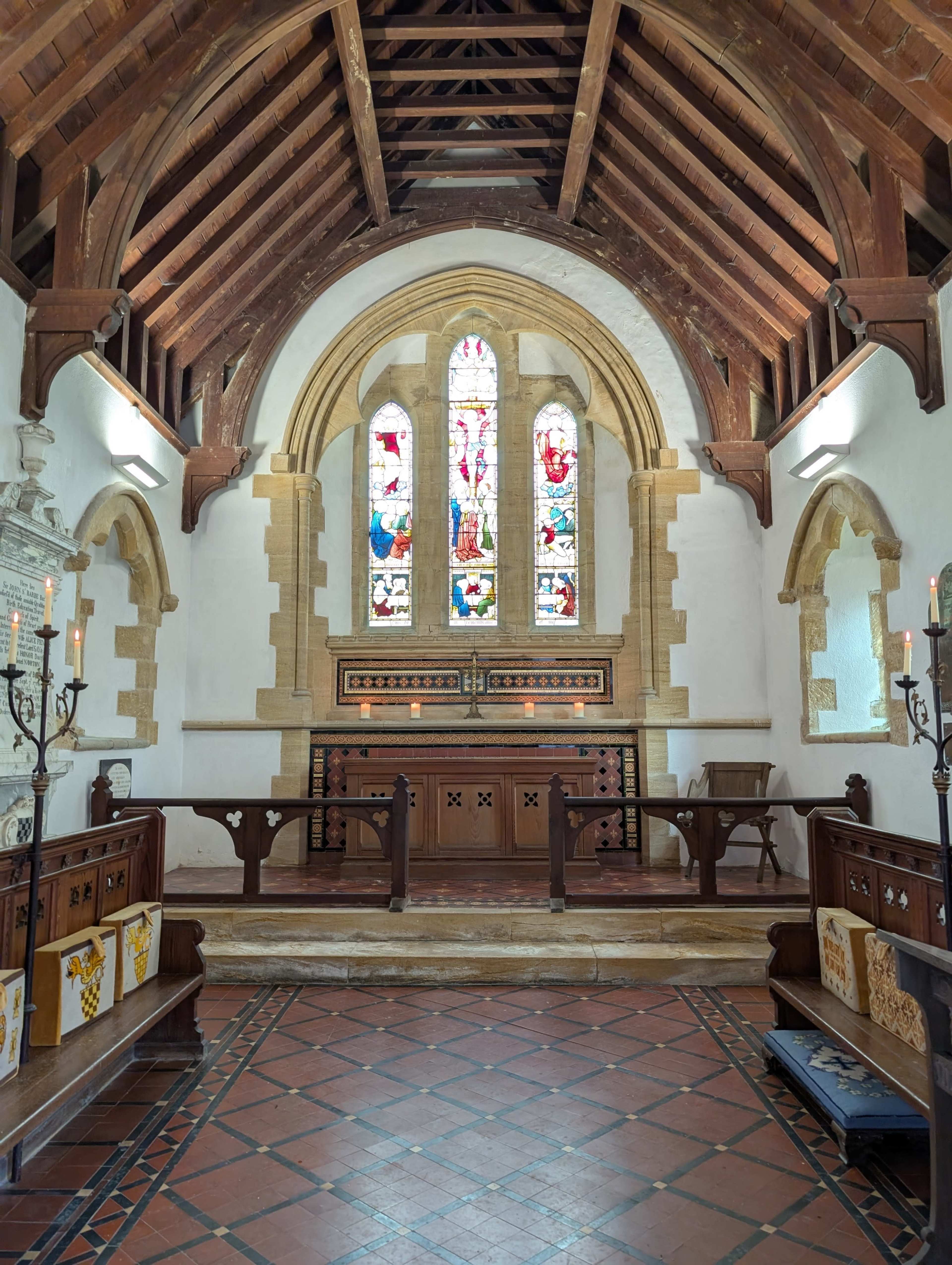 The interior of a small chapel features wooden beams, stained glass windows, and wooden pews arranged facing a raised altar.