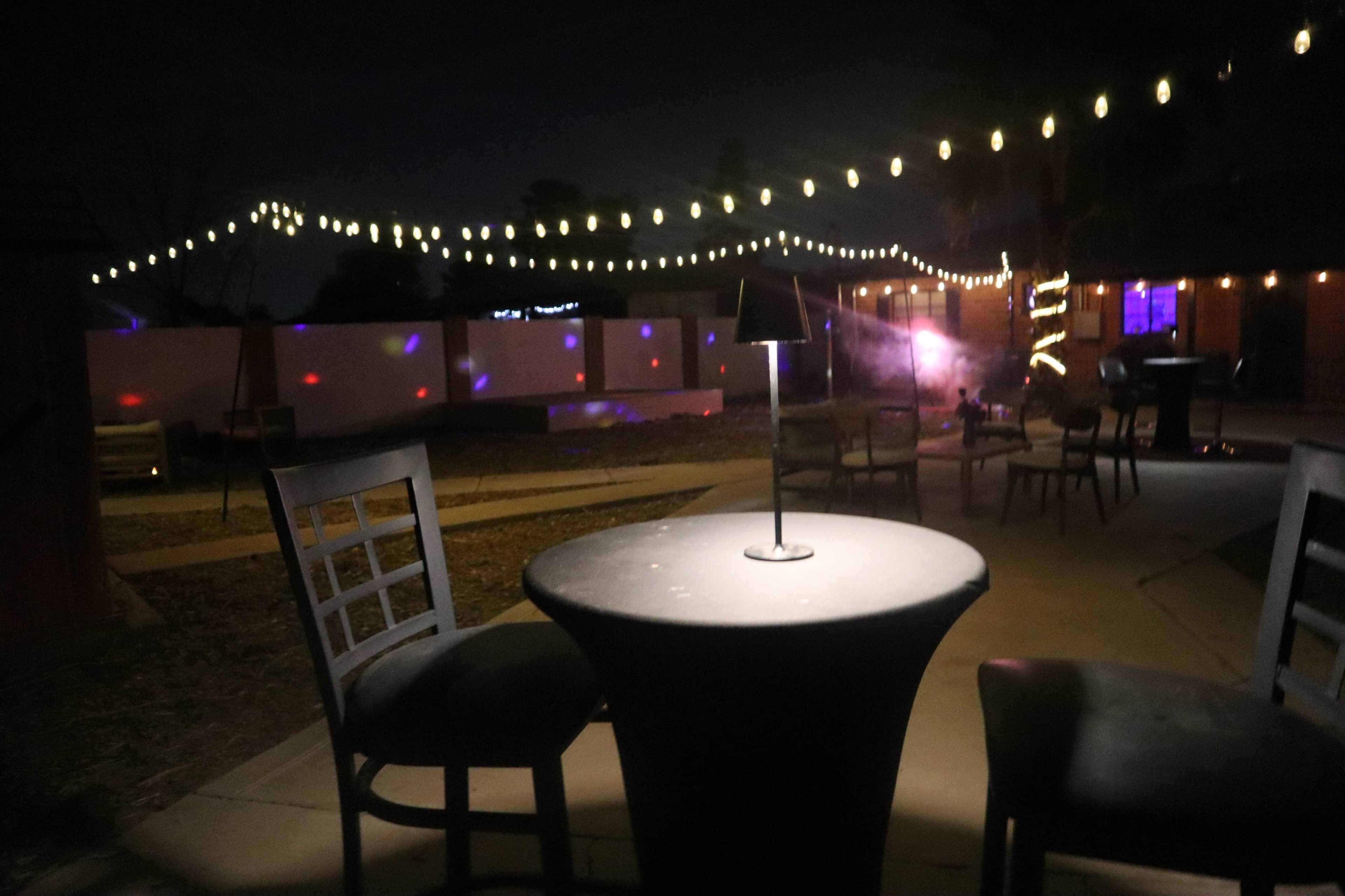 A table with a lamp stands in a dimly lit outdoor space adorned with string lights and scattered chairs.