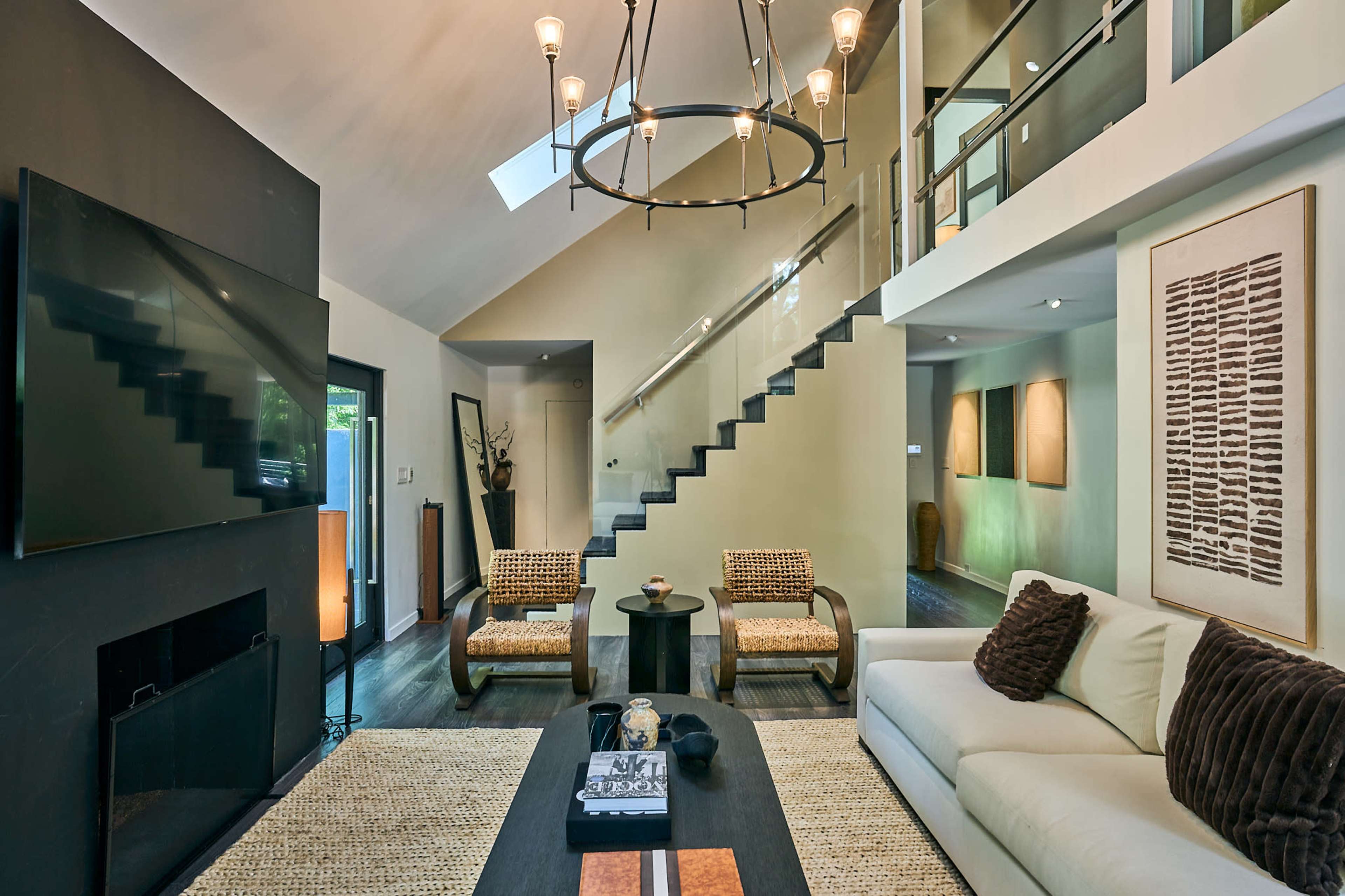 The image shows a modern living room with a high ceiling, large windows, a staircase, and contemporary furniture.