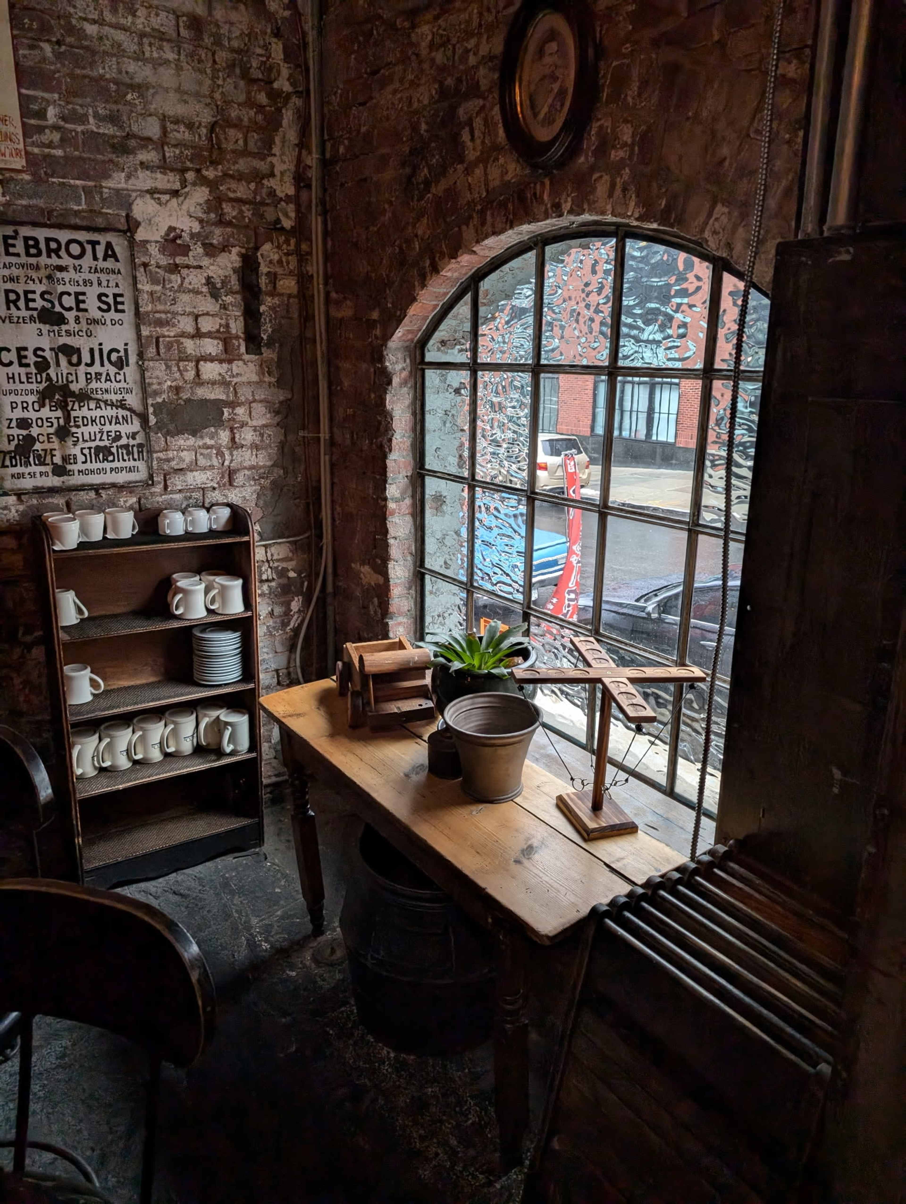 The image shows a rustic interior of a cafe with exposed brick walls, a wooden table, a plant, and a large window displaying a street outside.