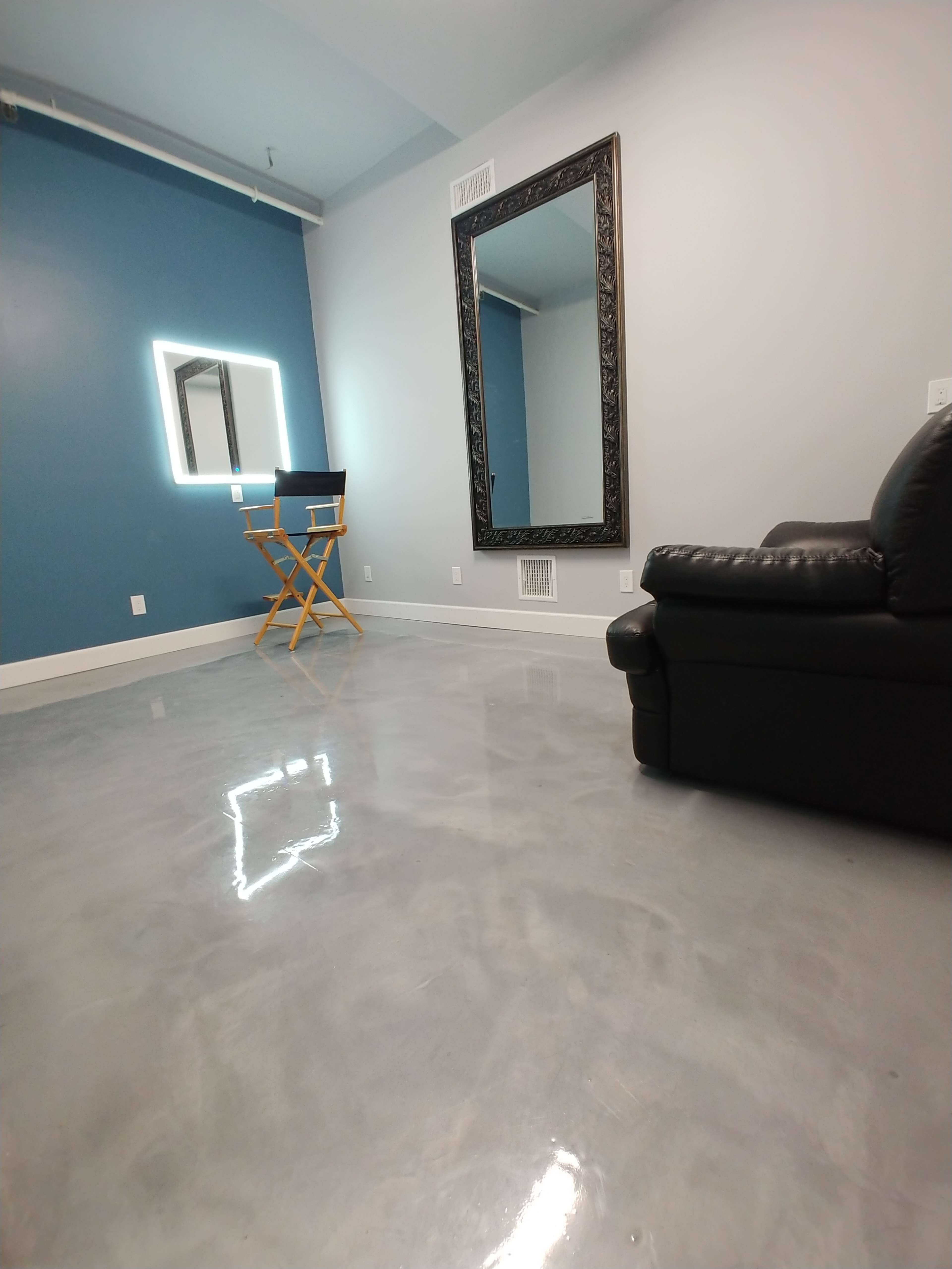 The image shows a modern room with a polished concrete floor, a blue accent wall, a large mirror, a chair positioned in front of a lighted mirror, and a black couch.