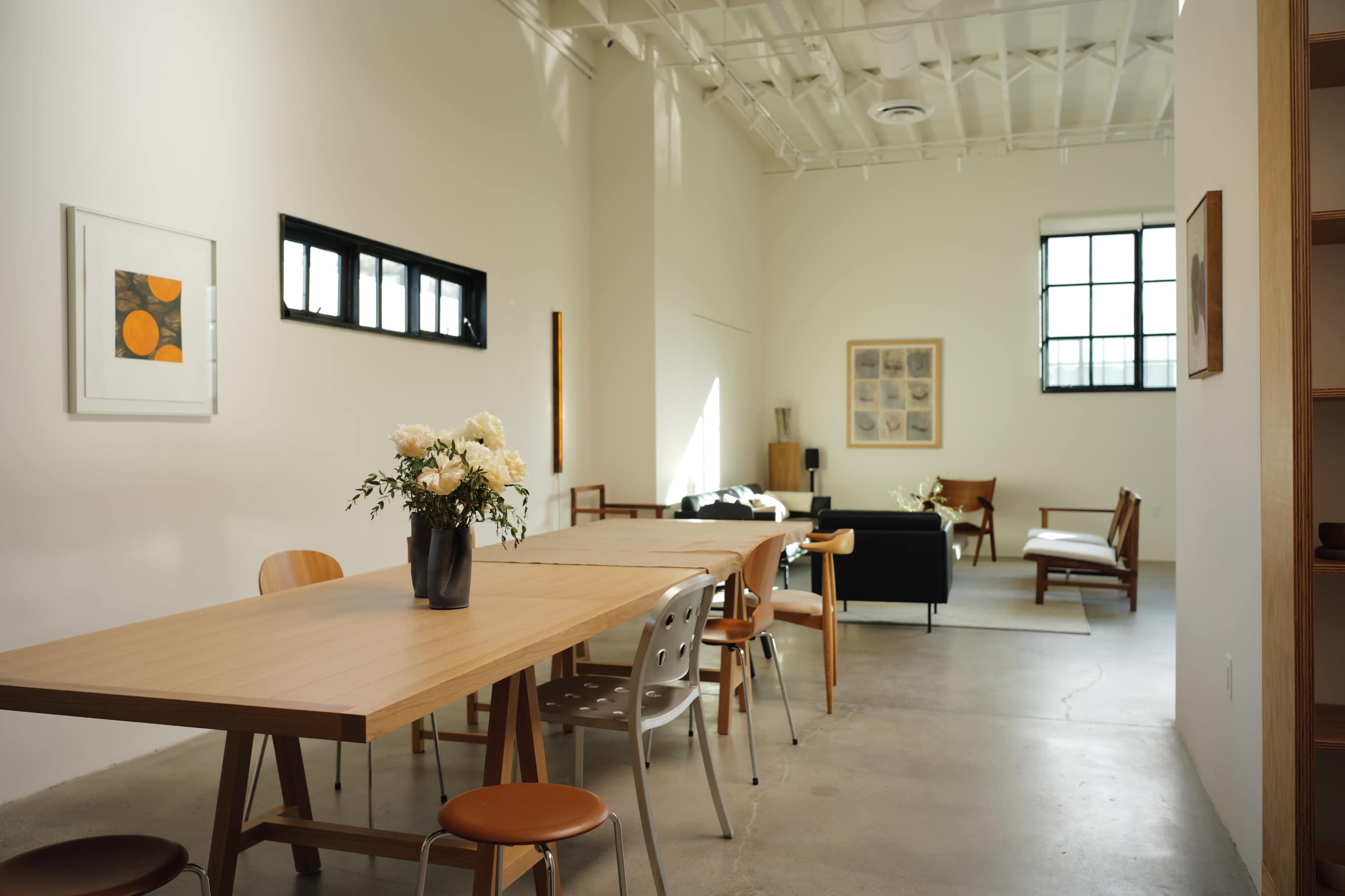 A spacious, minimalist room features a large wooden table with chairs, a flower arrangement in a vase, and various seating areas plus artwork on the walls.
