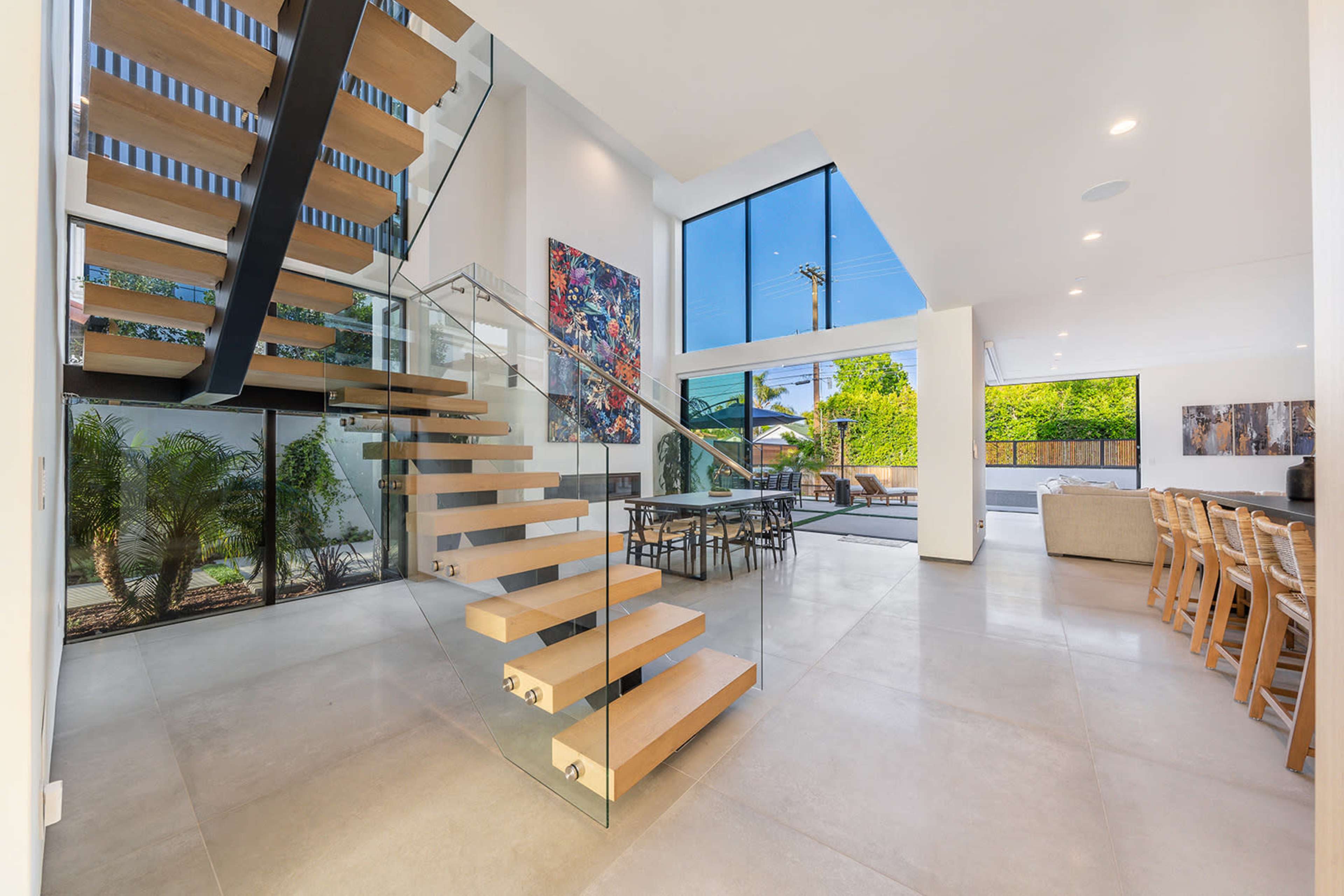A modern interior space featuring a floating staircase with wooden steps, large windows providing natural light, and a dining area with a table and chairs.