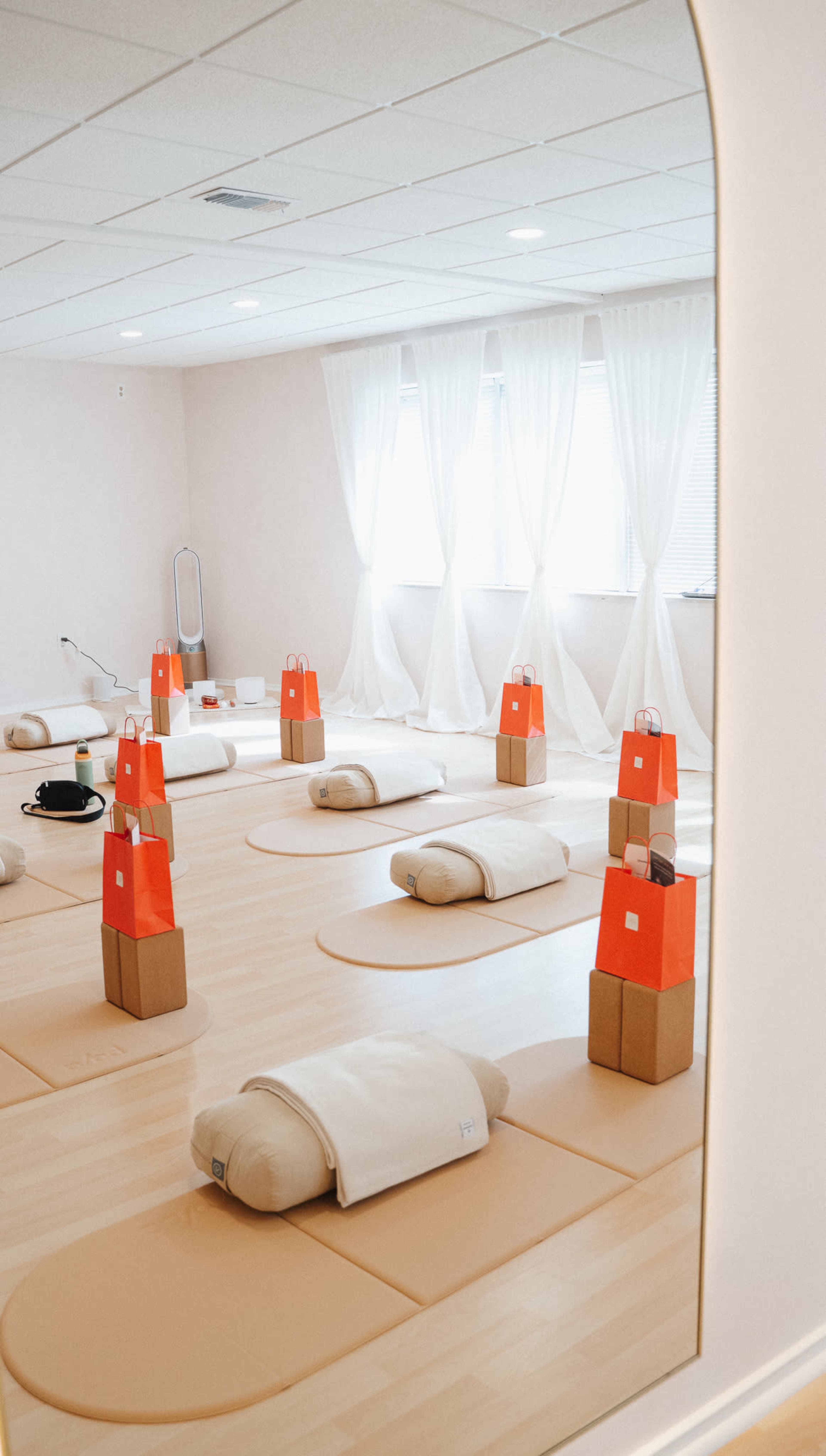 The image shows a spacious, well-lit yoga studio featuring several yoga mats arranged on the floor, each with a stack of cushions and small bags on top.