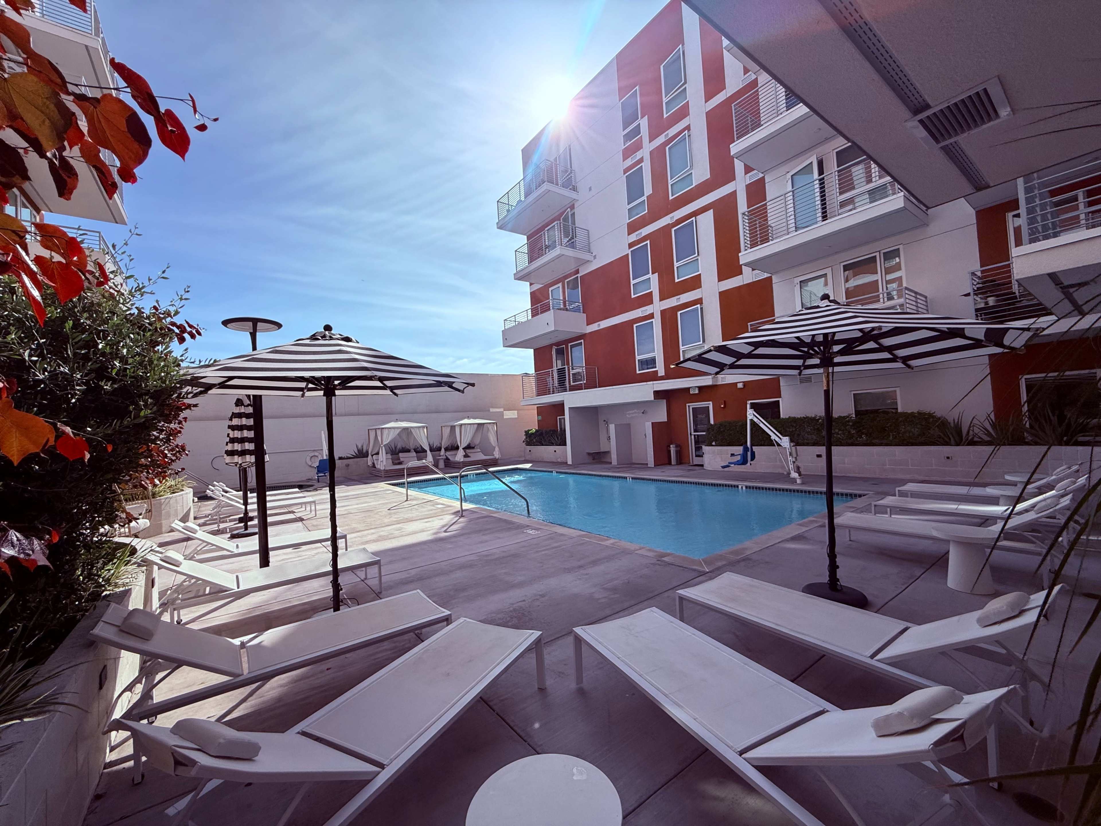 The image shows a modern pool area surrounded by lounge chairs and umbrellas, with a building featuring orange and white facades in the background.