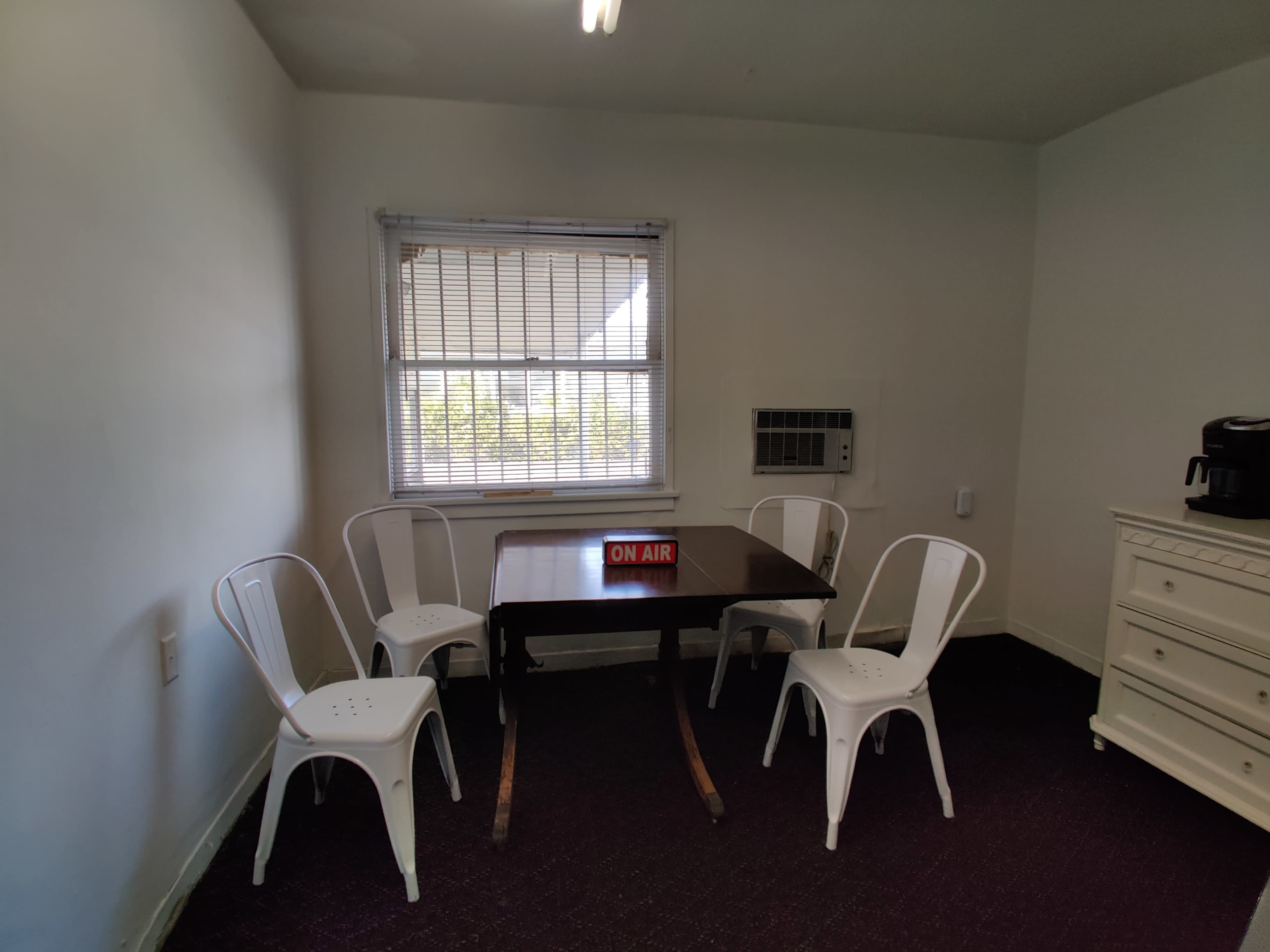 A small room features a table with four white chairs, a window with blinds, and an "ON AIR" sign.
