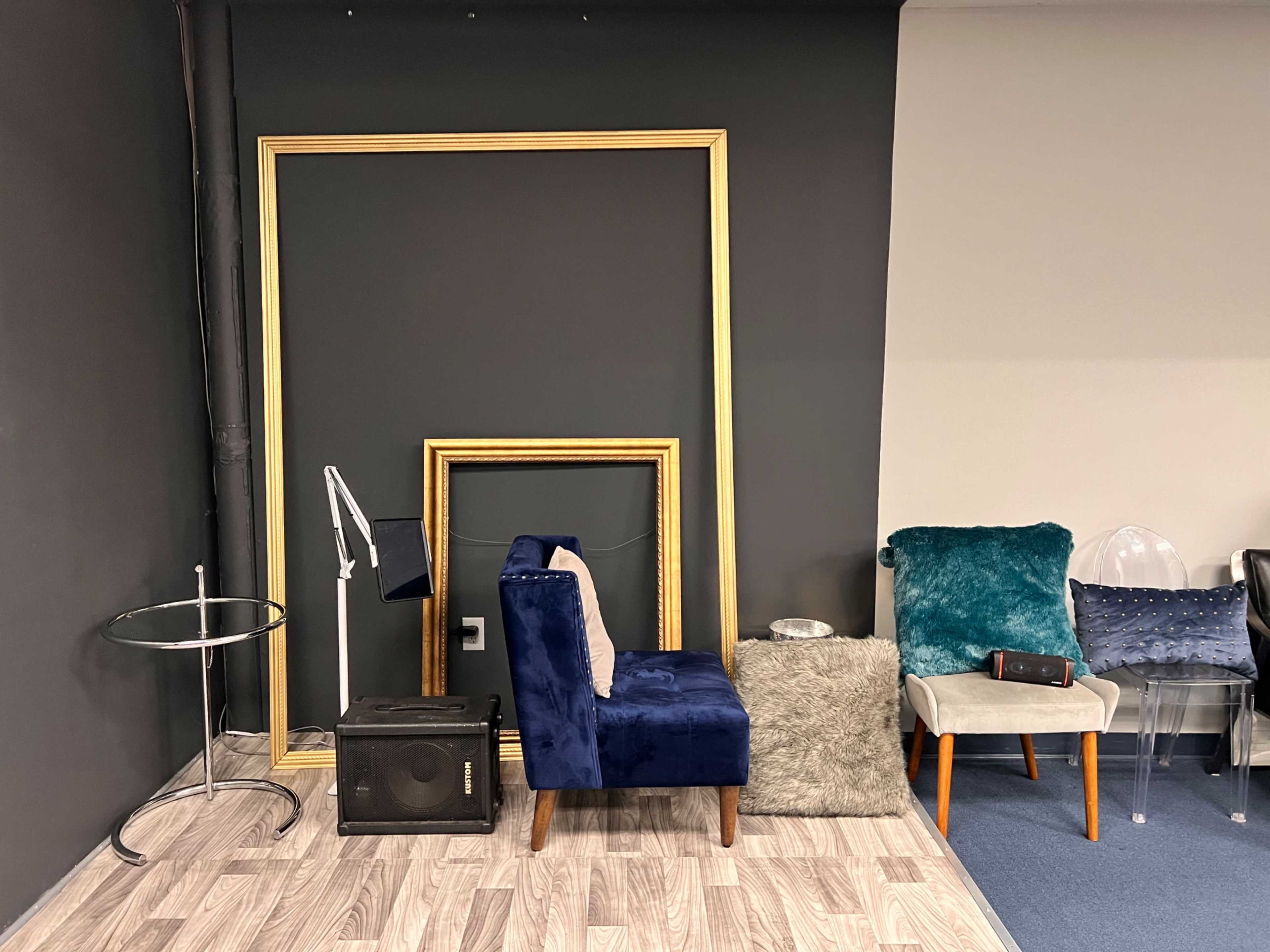 A room corner with two empty picture frames, a blue upholstered chair, a circular glass table, a lamp, and scattered cushions.