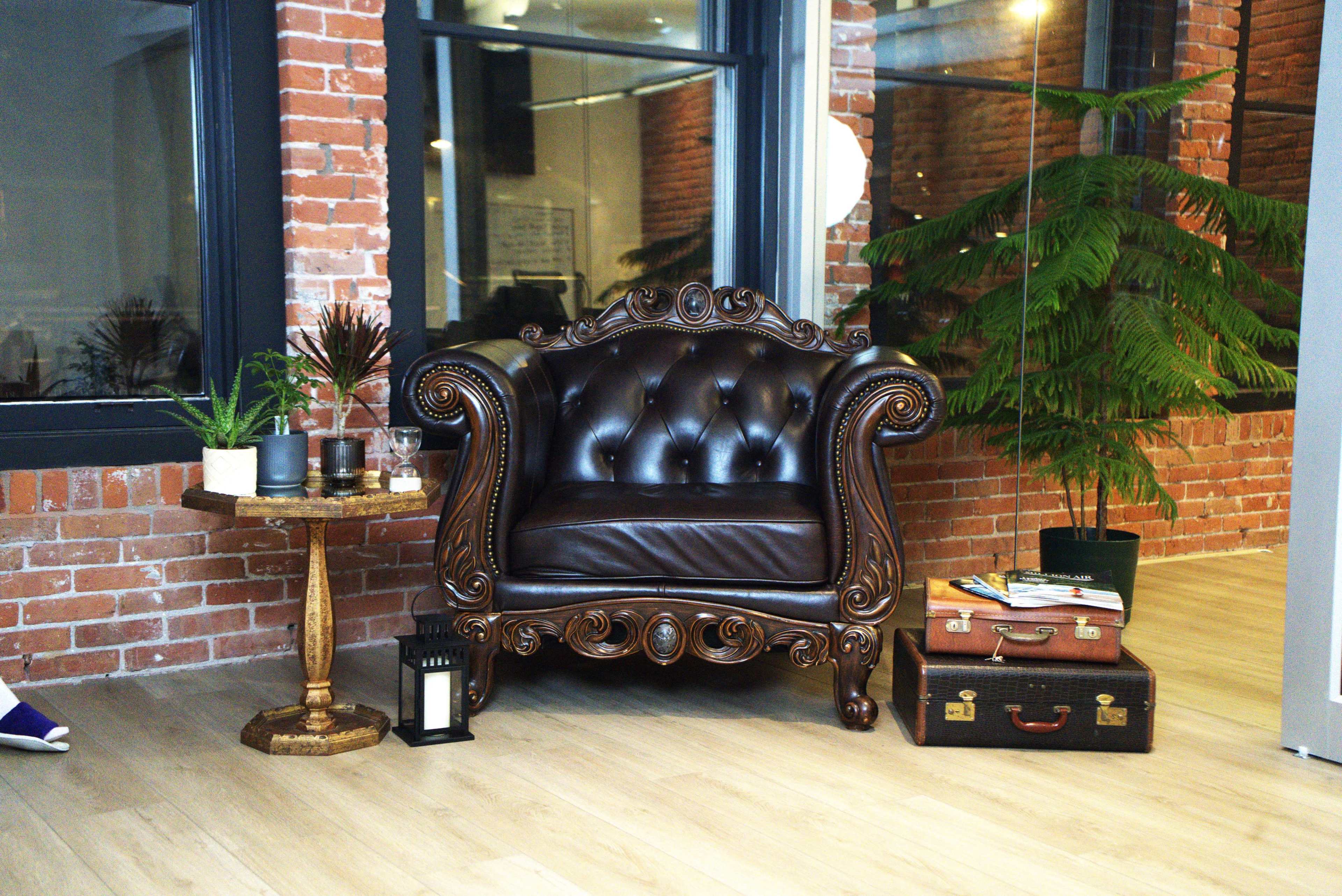A vintage-style brown leather armchair is placed next to a small round wooden table, surrounded by potted plants in a modern brick-walled room.