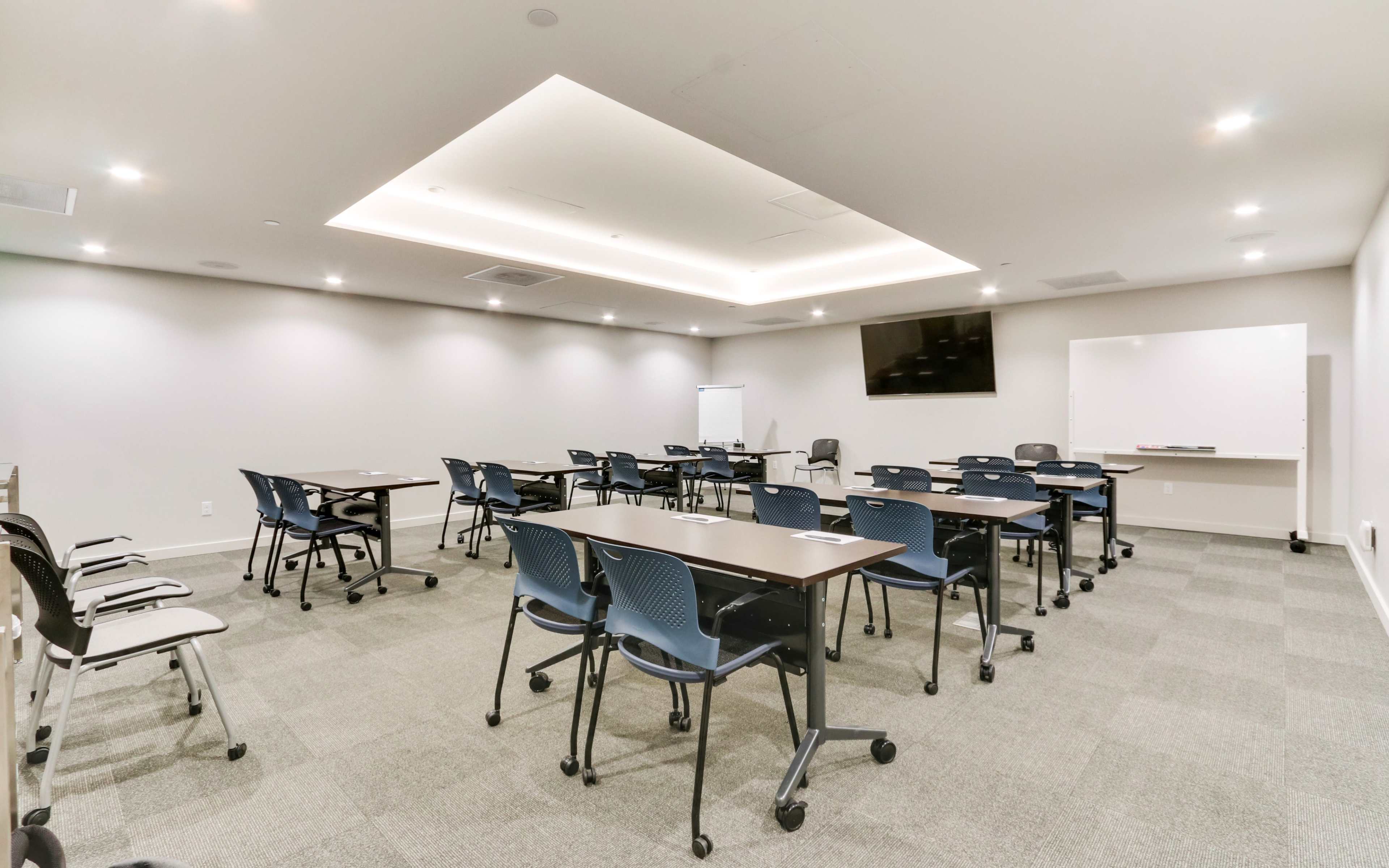 A spacious meeting room with several rows of tables and chairs arranged for a seminar, featuring a large screen and whiteboard.