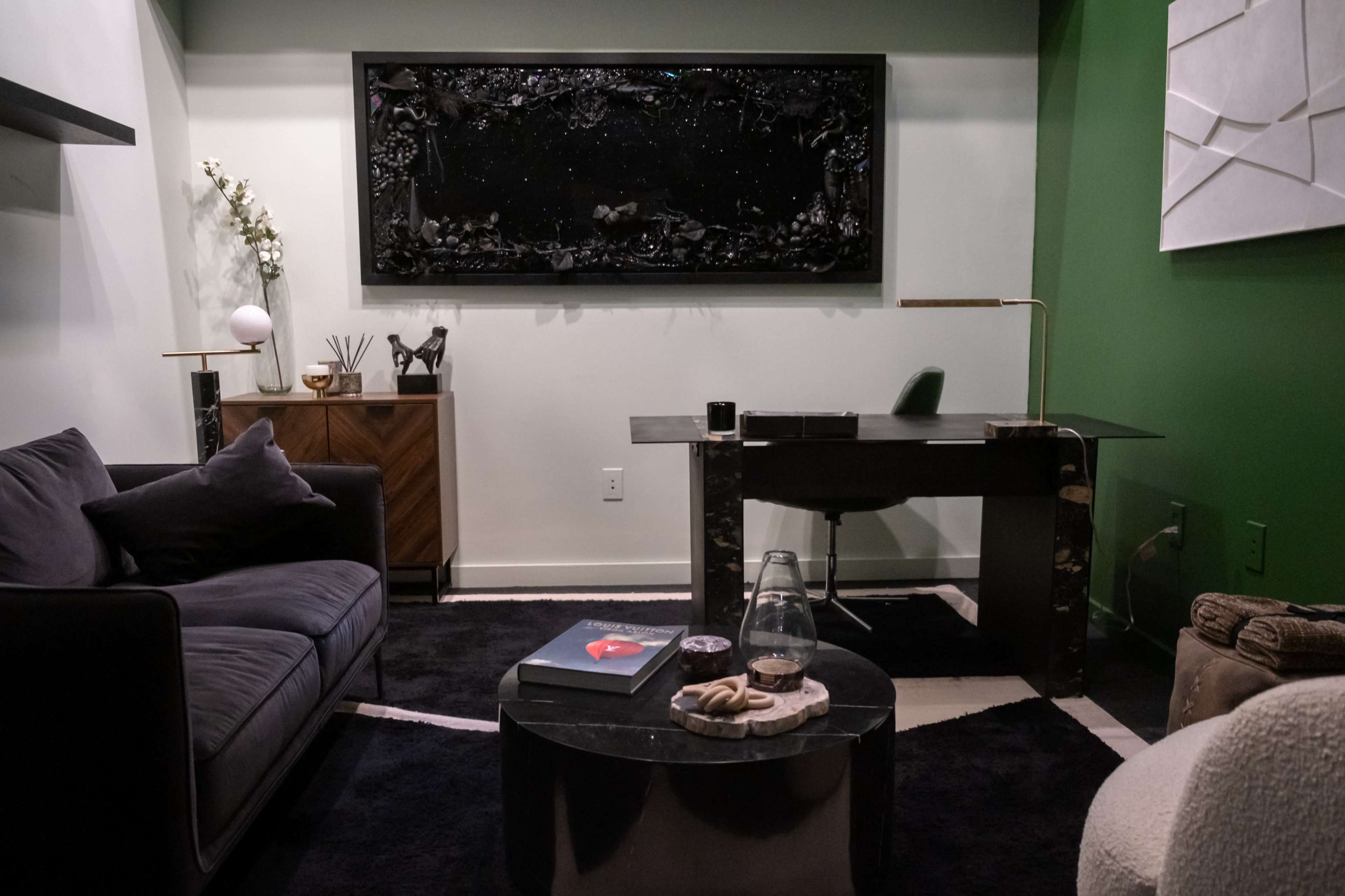 The image shows a modern office space featuring a dark wood desk, a black sofa, and a circular coffee table, with green and white walls and decorative artwork.