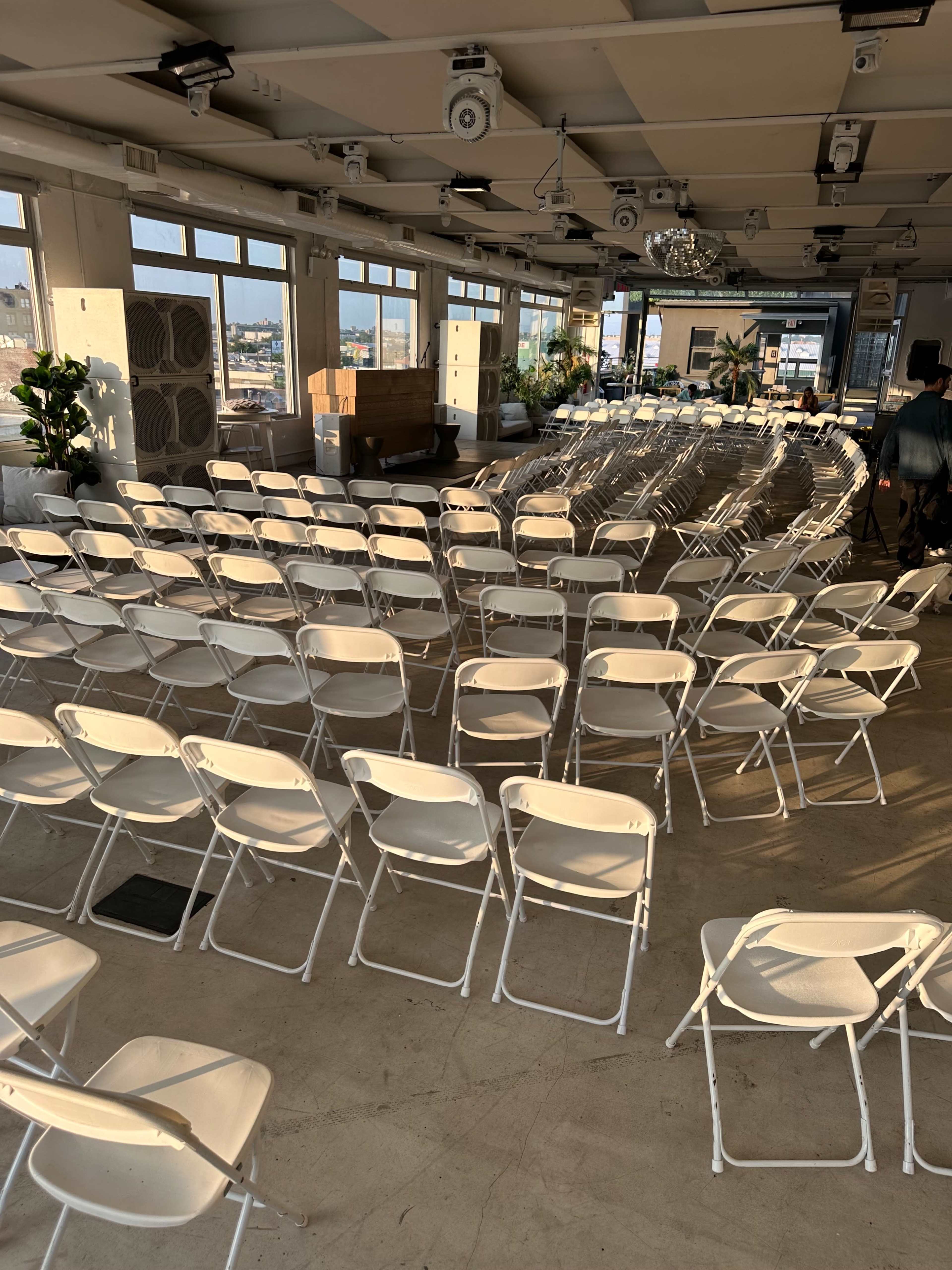 The image shows a spacious venue with rows of empty white folding chairs arranged in a circular pattern, large windows revealing a view of the outdoors.