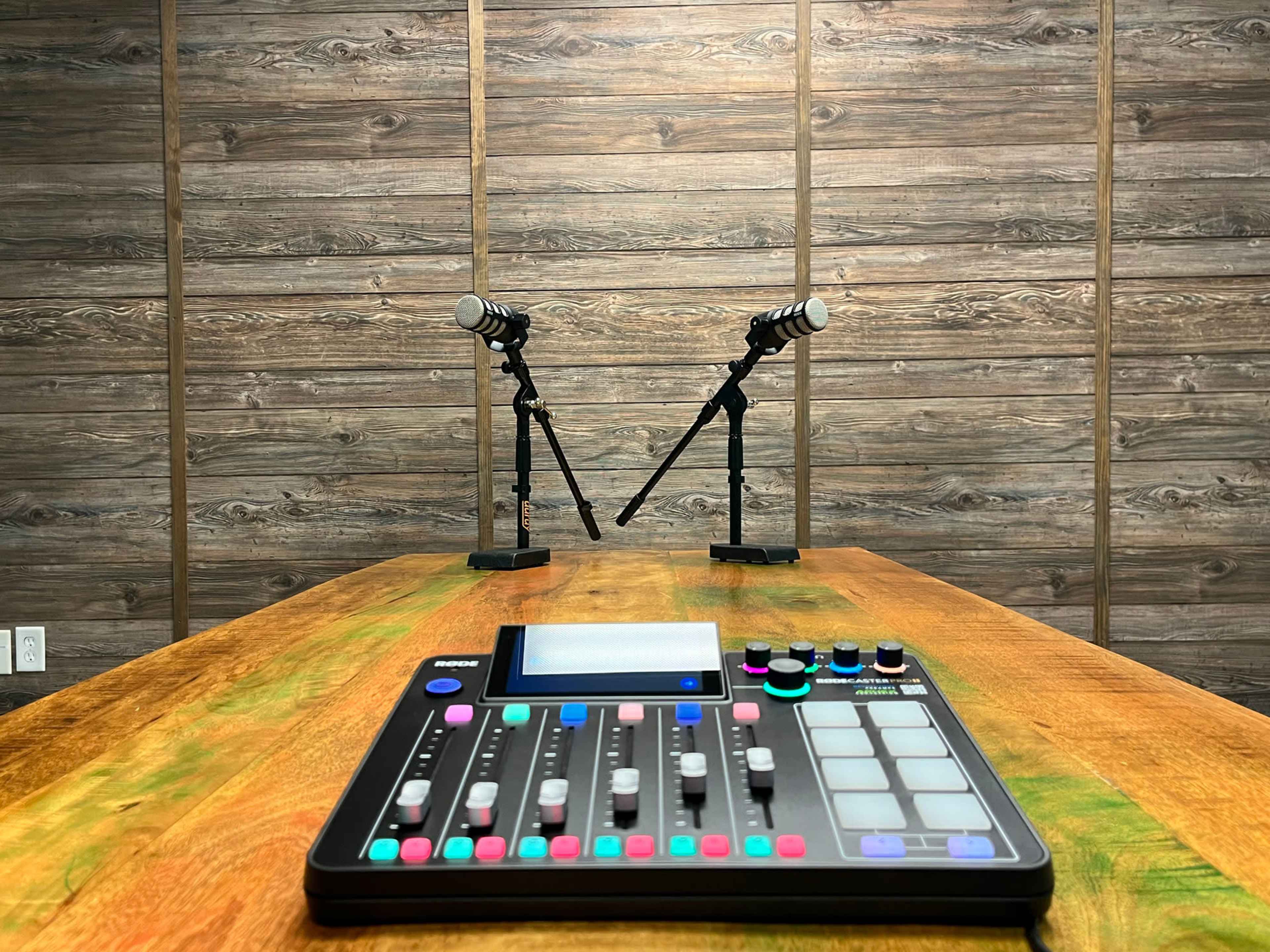 A wooden table with a digital audio mixer in the foreground and two microphones on stands set up against a wooden wall.
