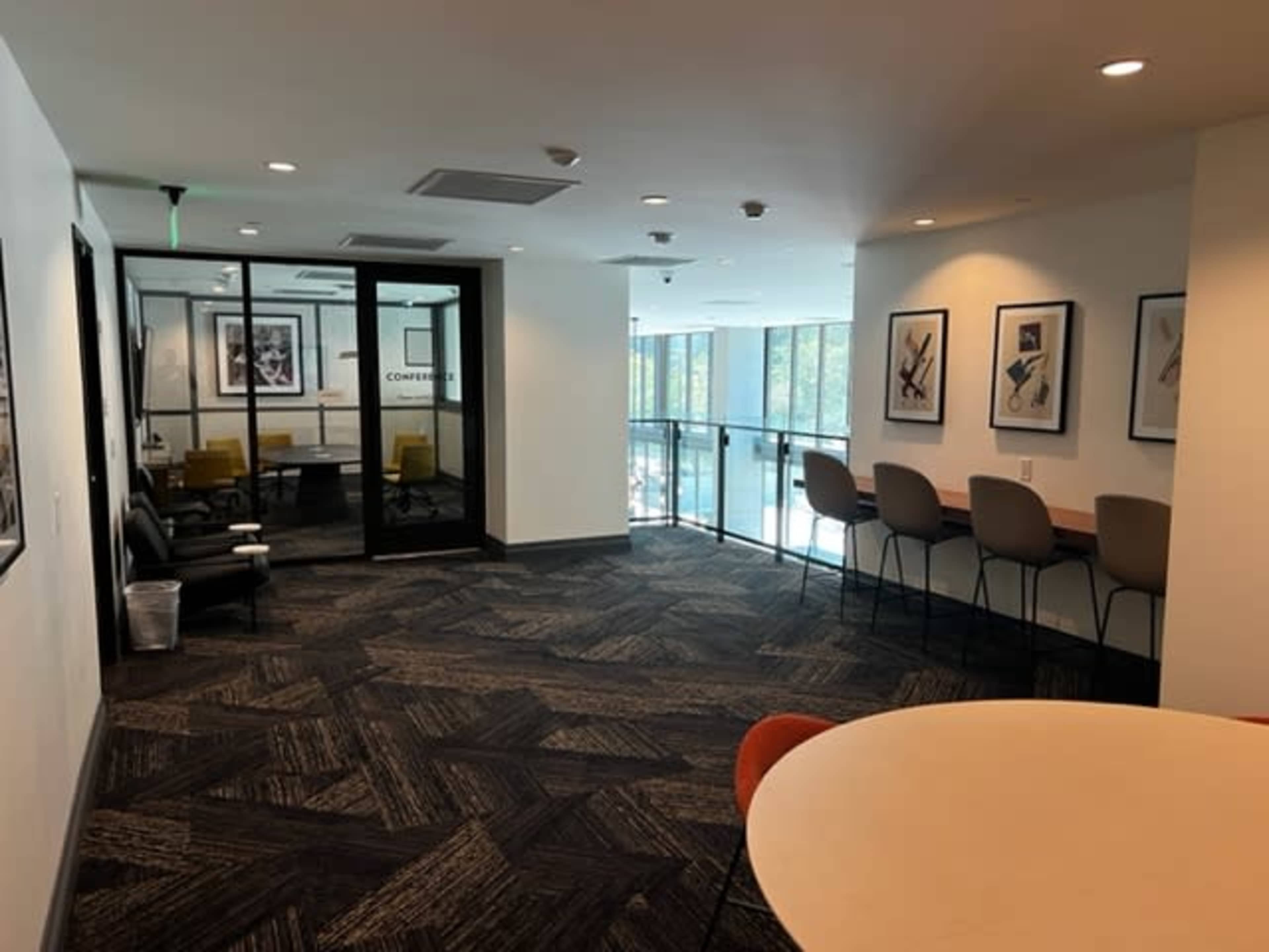 The image shows a modern office corridor with seating areas, framed artwork on the walls, and a glass railing overlooking a bright space.