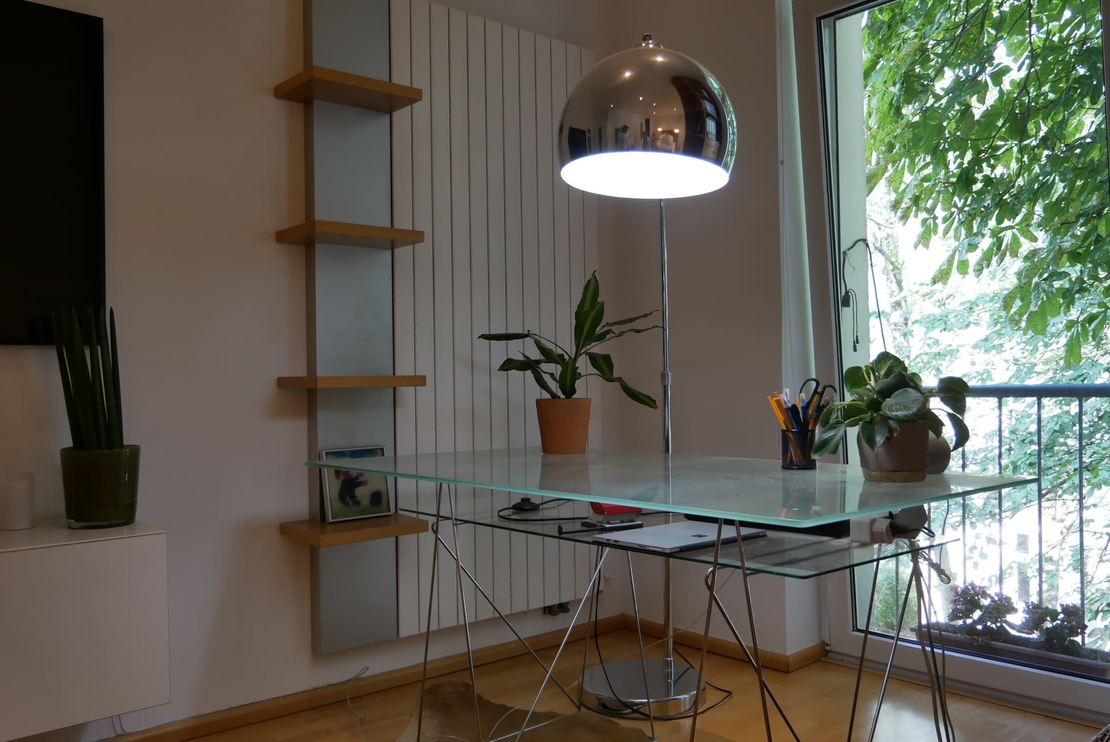 A modern glass desk is positioned next to a window with plants and a stylish lamp on top.