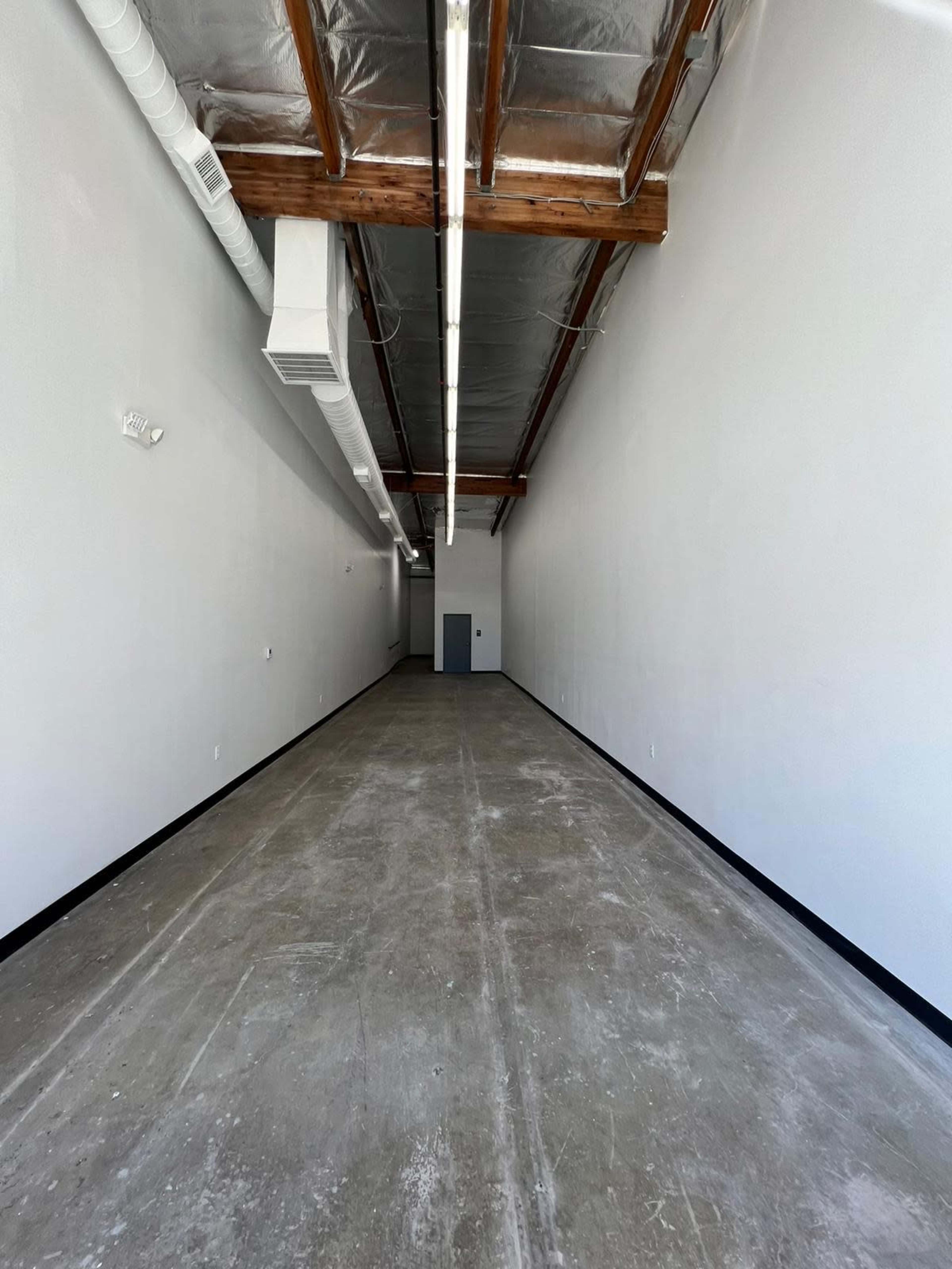 The image shows a long, empty hallway with a concrete floor and exposed wooden beams on the ceiling.