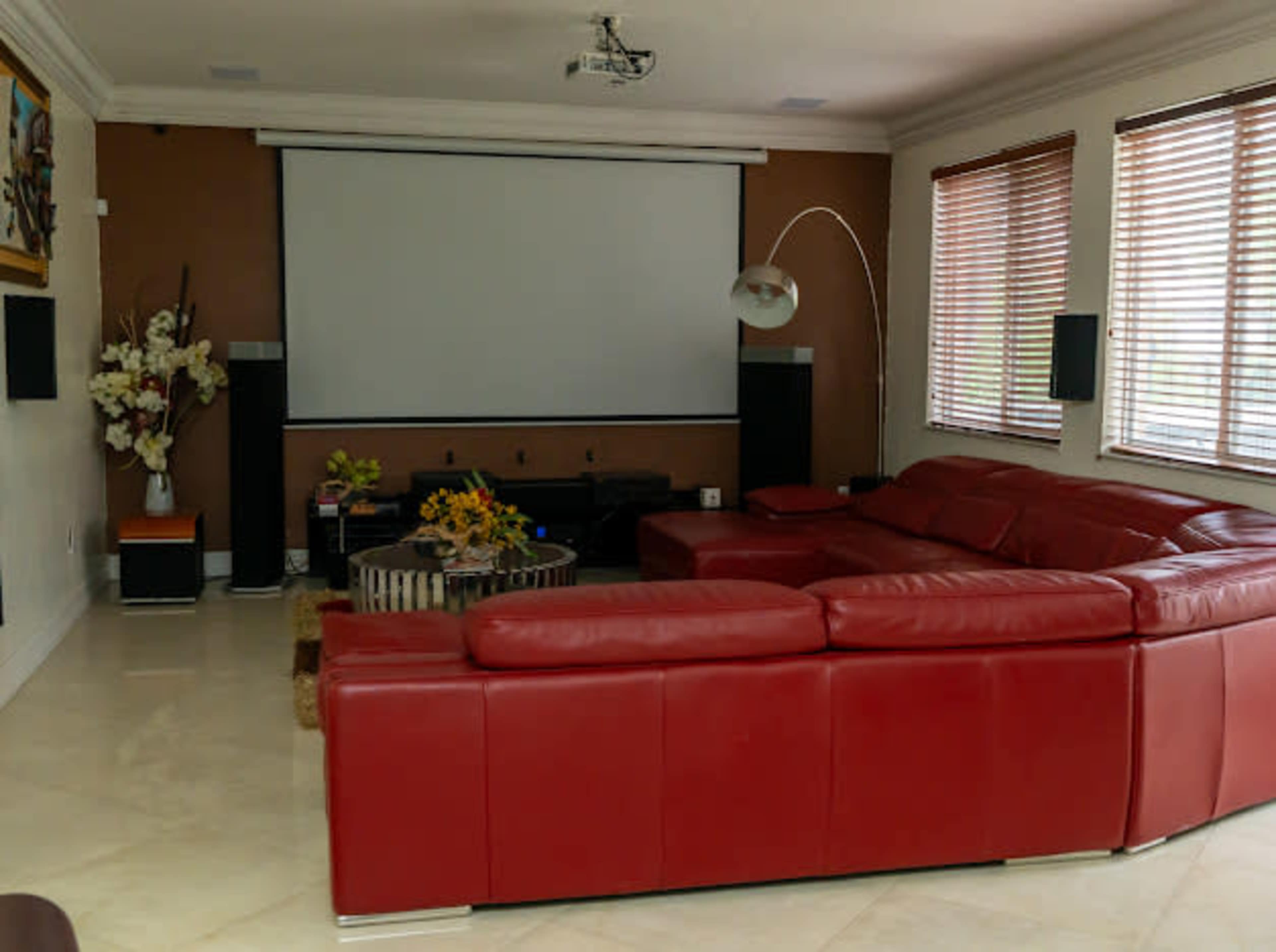 A spacious living room featuring a large red sectional sofa, a flat-screen projector screen, and natural light coming through wooden blinds.