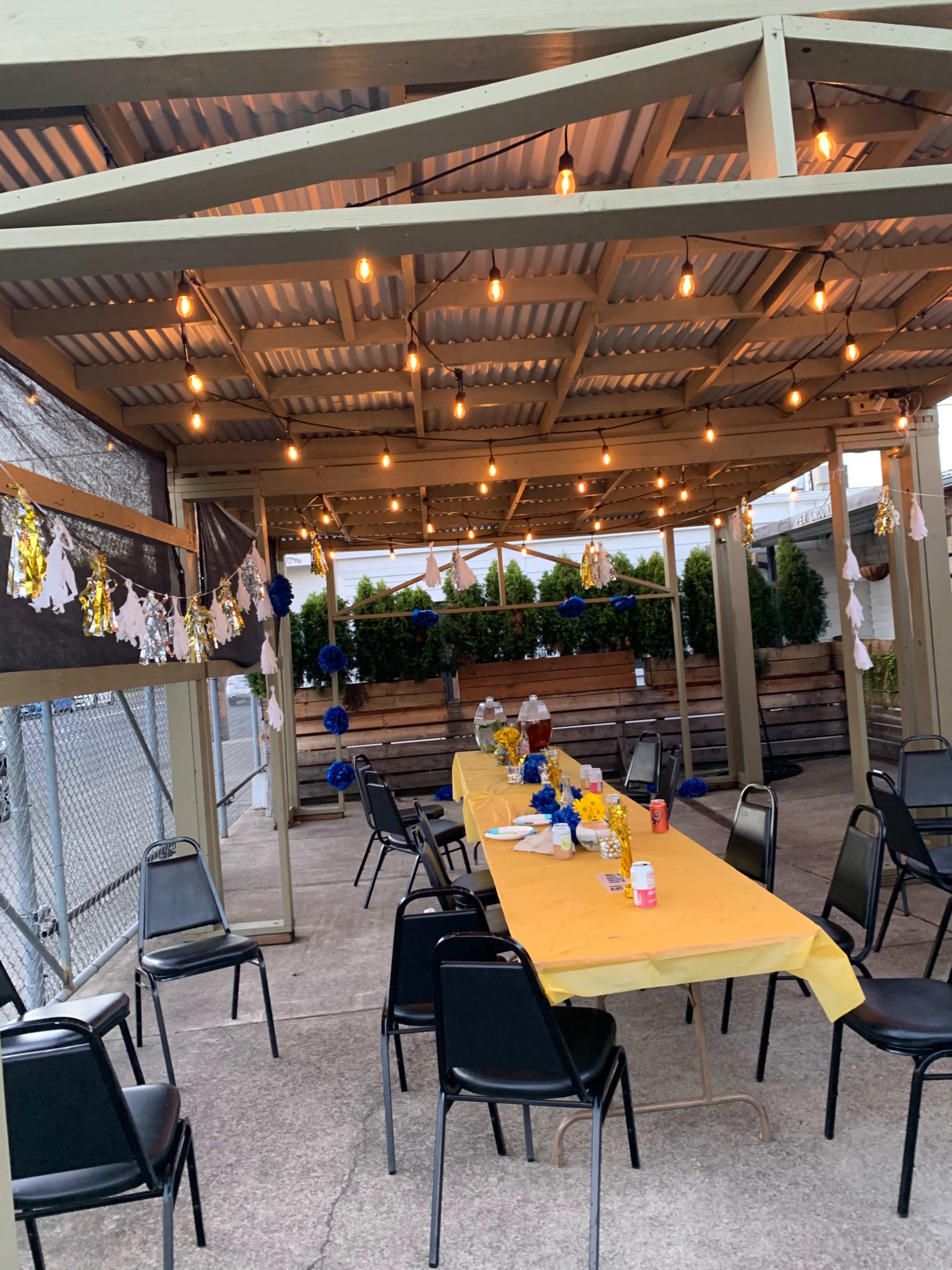 A decorated outdoor space features a long table set with yellow cloth and floral arrangements, under string lights and banners.
