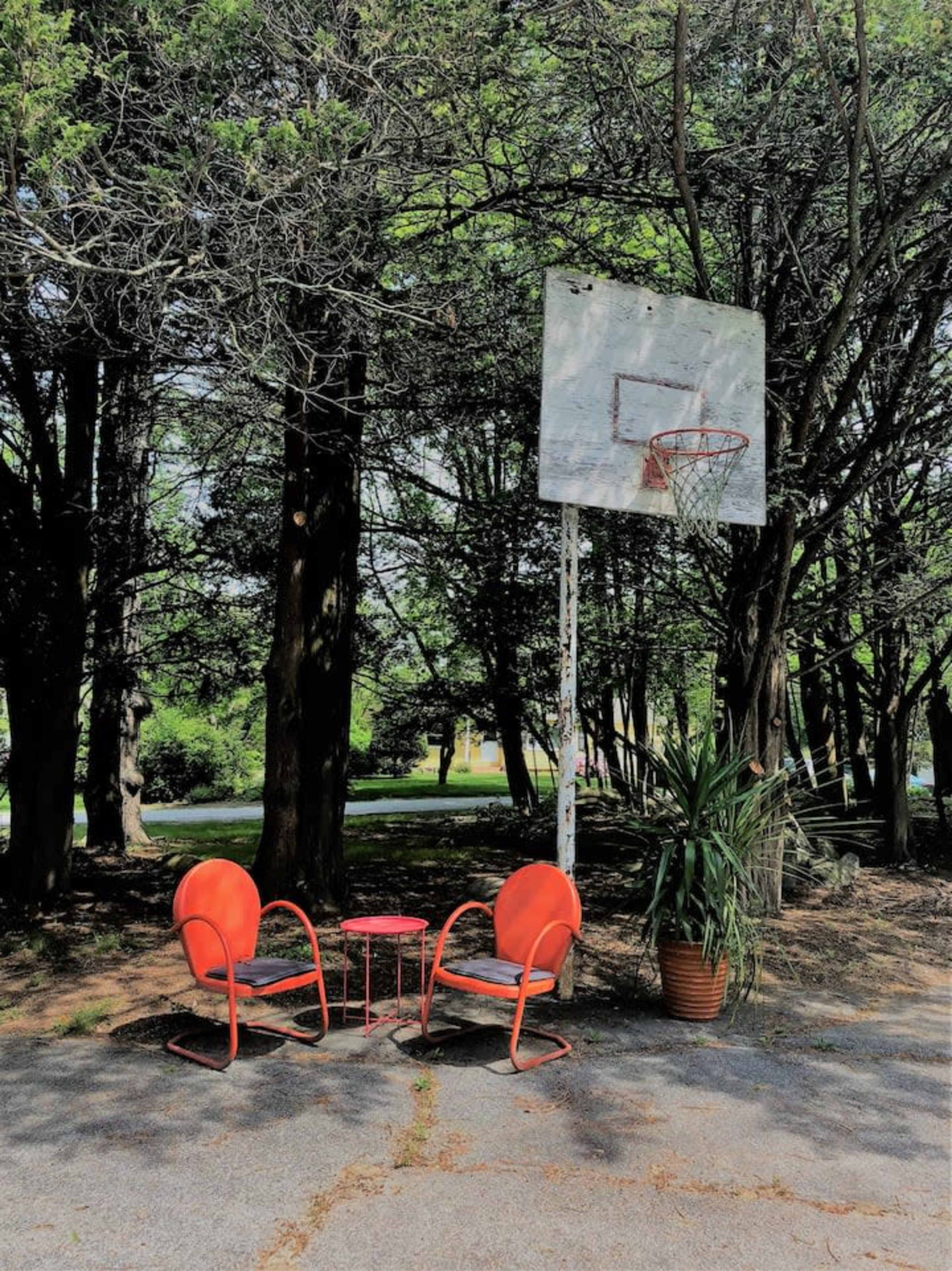 Two orange chairs and a small table are set up near a basketball hoop surrounded by trees.