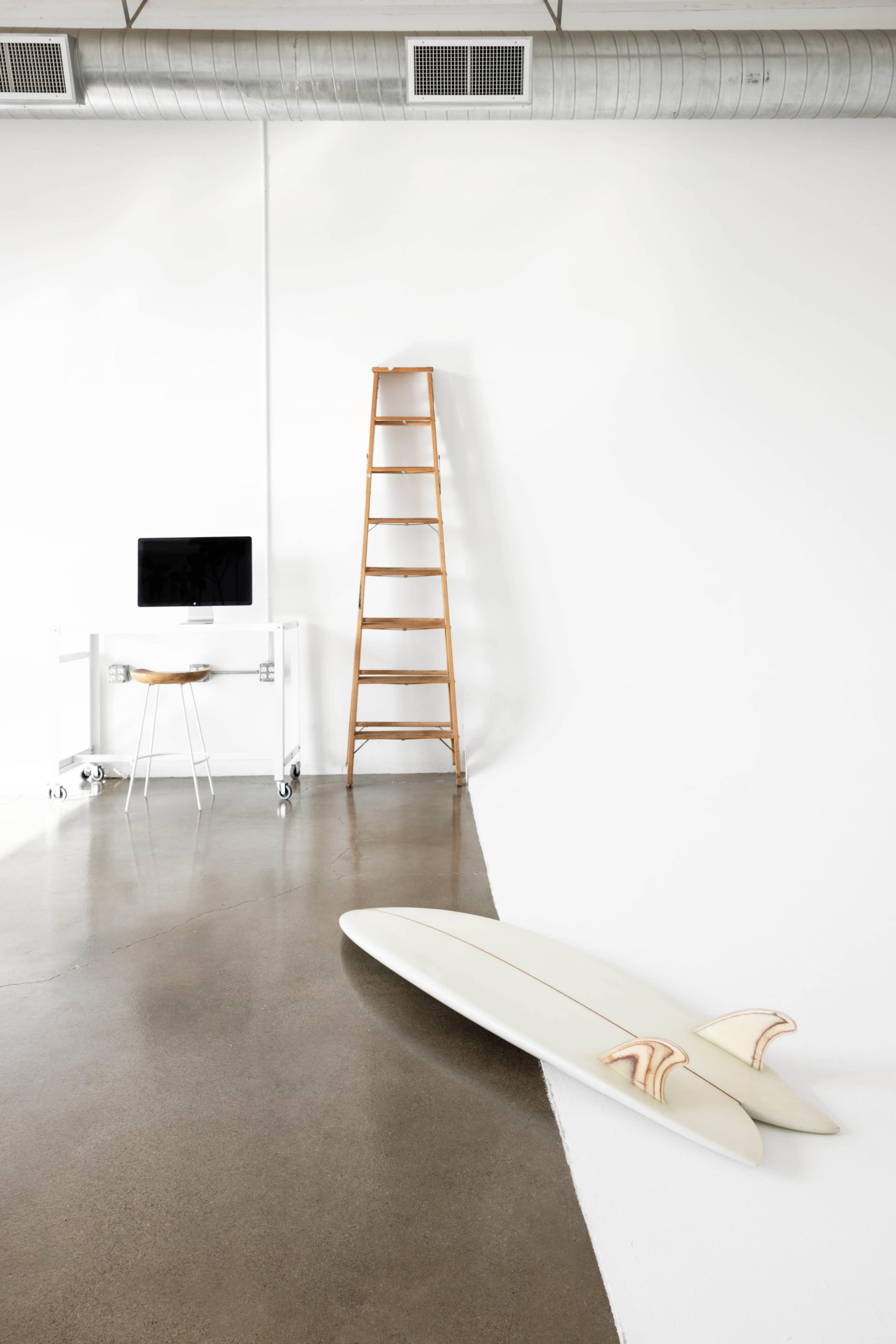 A minimalist interior featuring a computer desk, a wooden ladder against a wall, and two surfboards on a polished concrete floor.