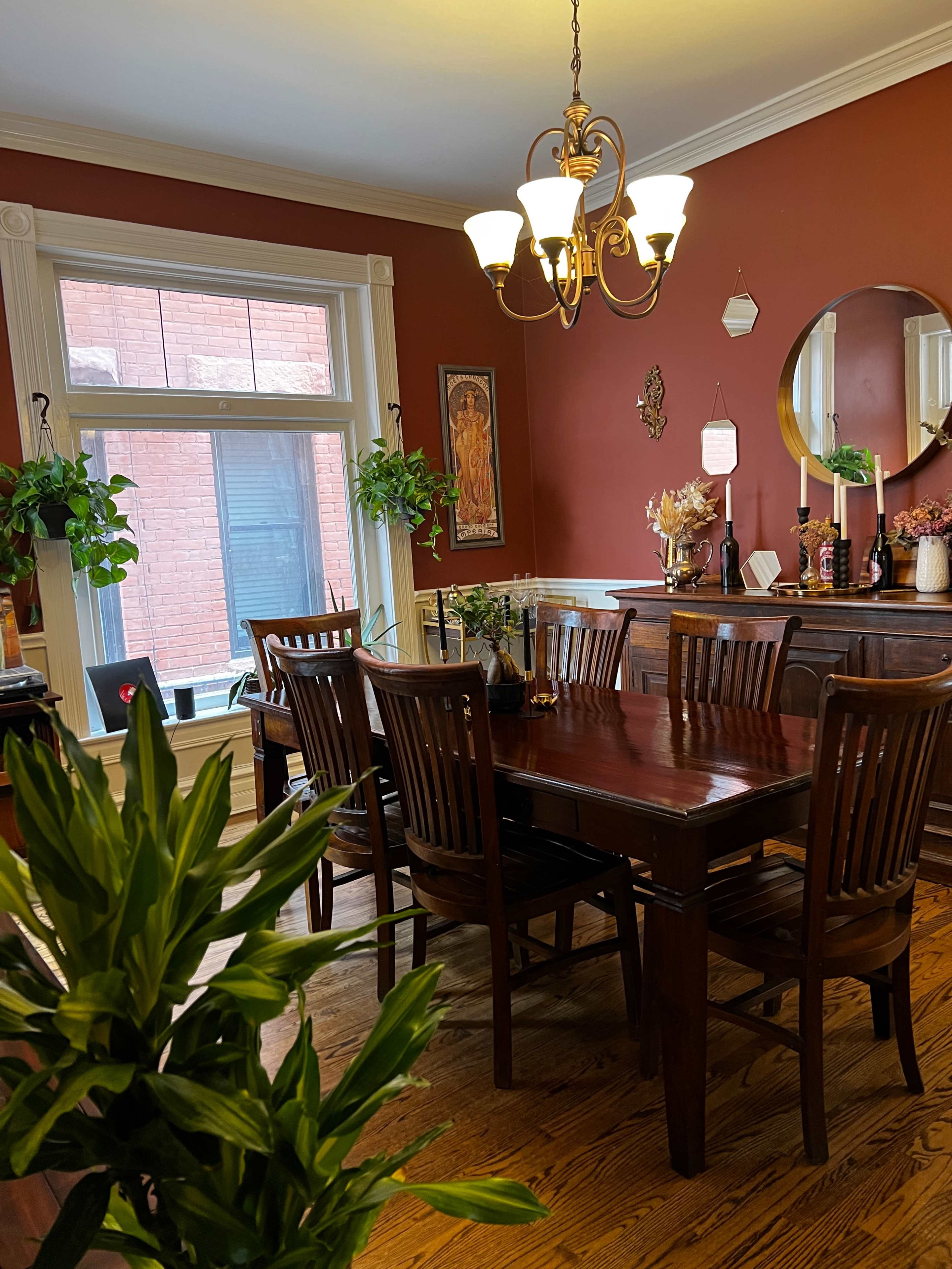 A wooden dining table is surrounded by chairs in a warmly colored room with plants, a chandelier, and a decorative mirror.