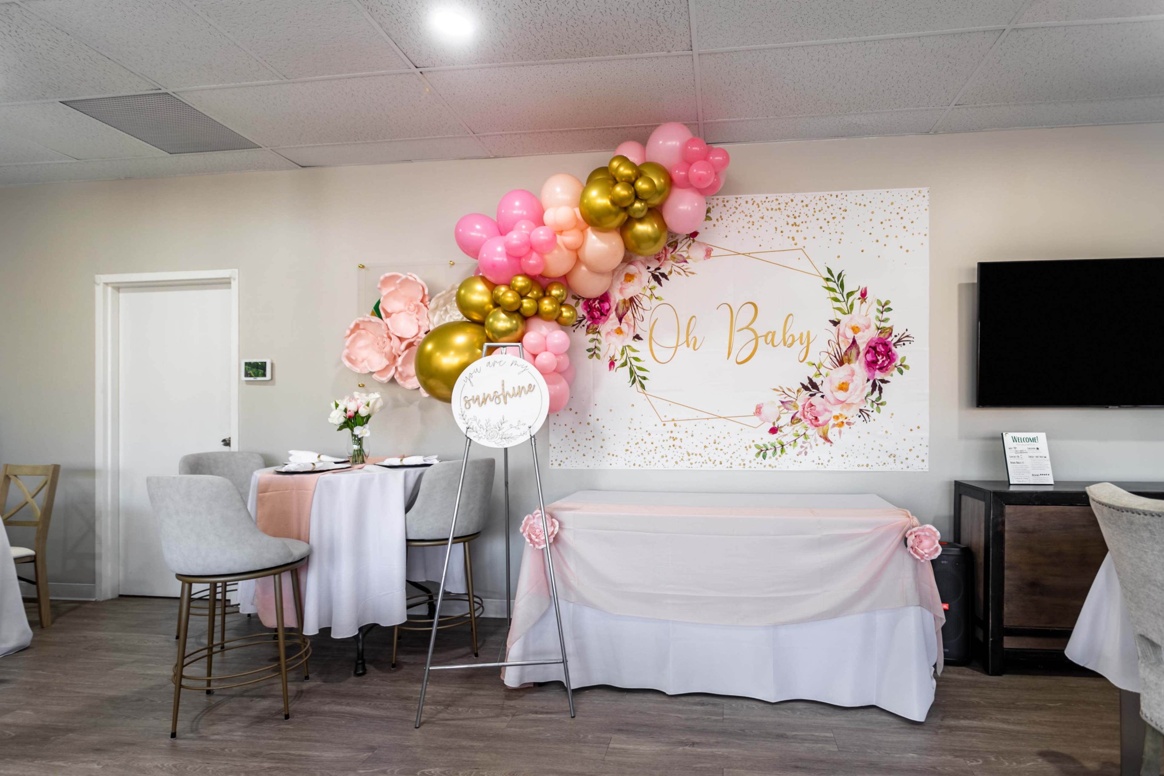 A decorated event space features a wall with a floral-themed "Oh Baby" sign, adorned with a colorful balloon arrangement and elegantly set tables.