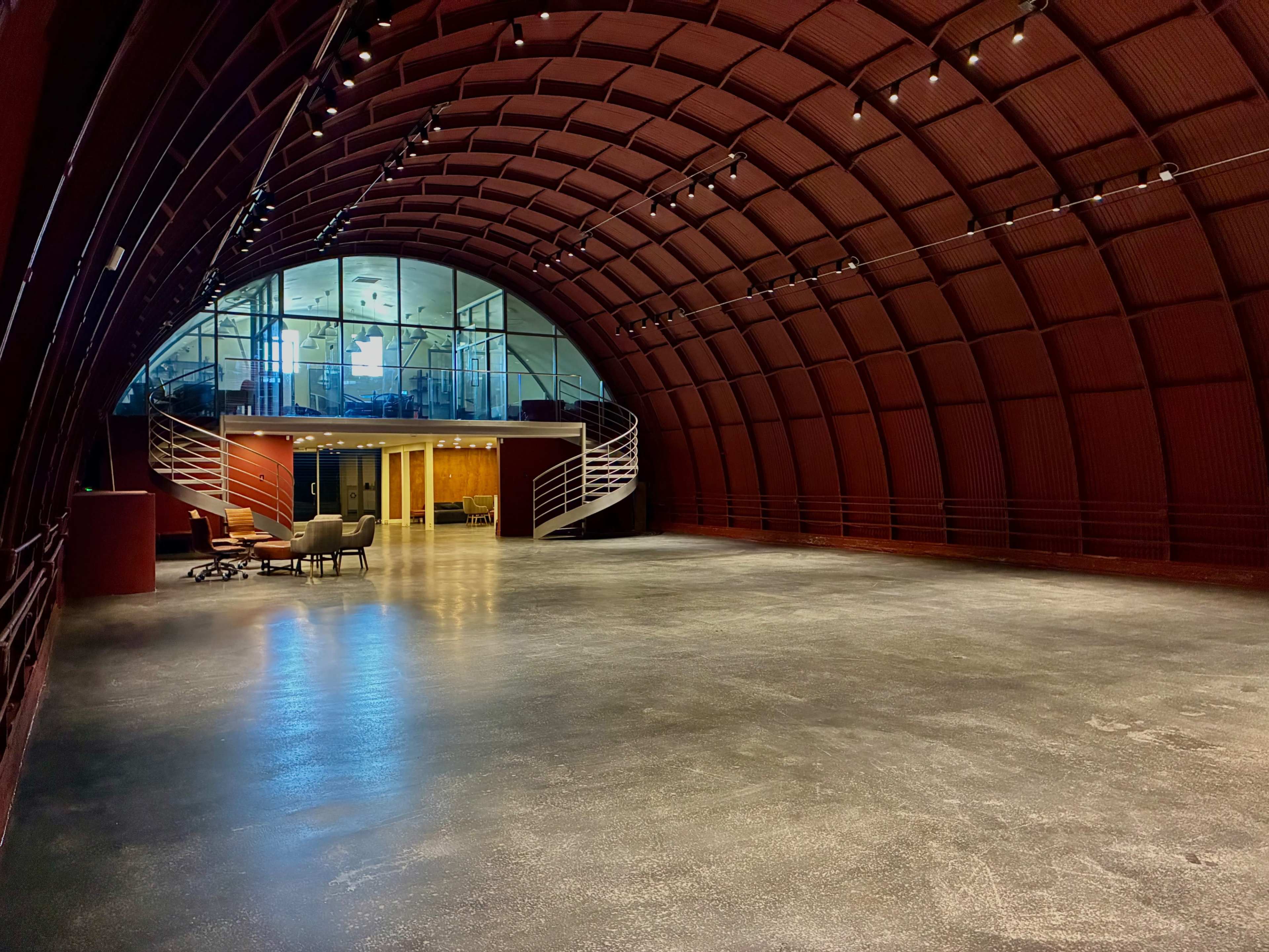 The image shows an expansive, modern interior space with a curved, red arching ceiling and a glass-walled area at one end.