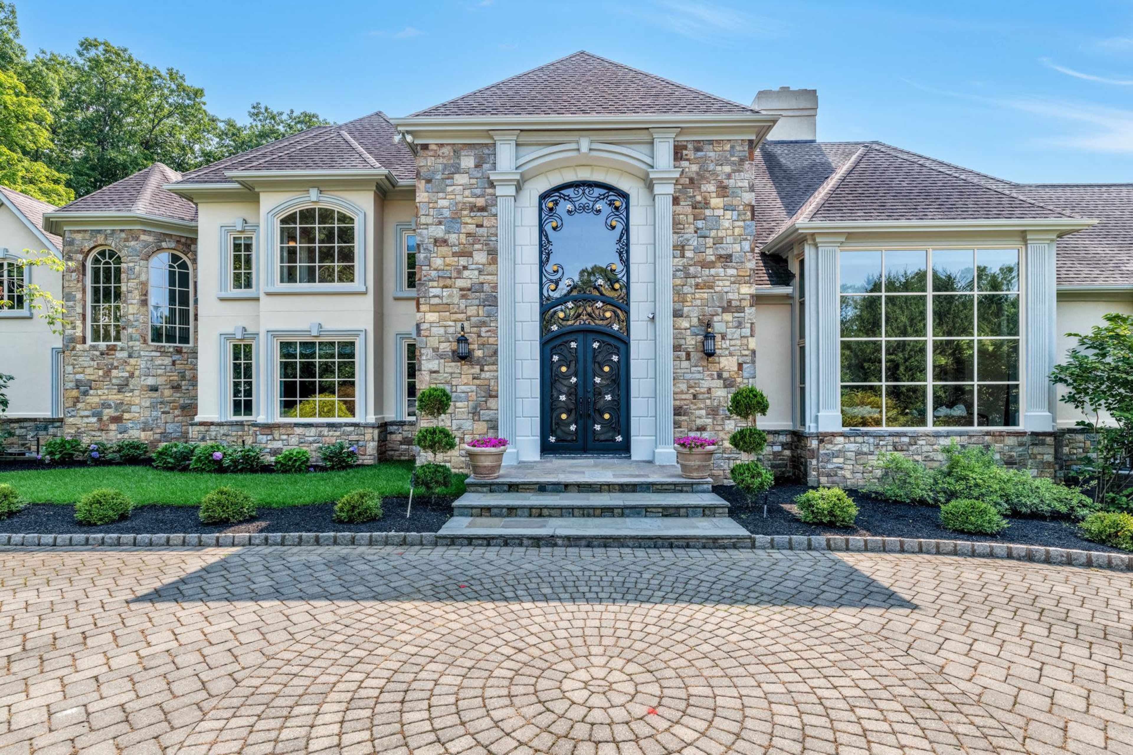The image shows a large, upscale home with a stone facade, prominent entryway, and landscaped front yard featuring round flower beds.