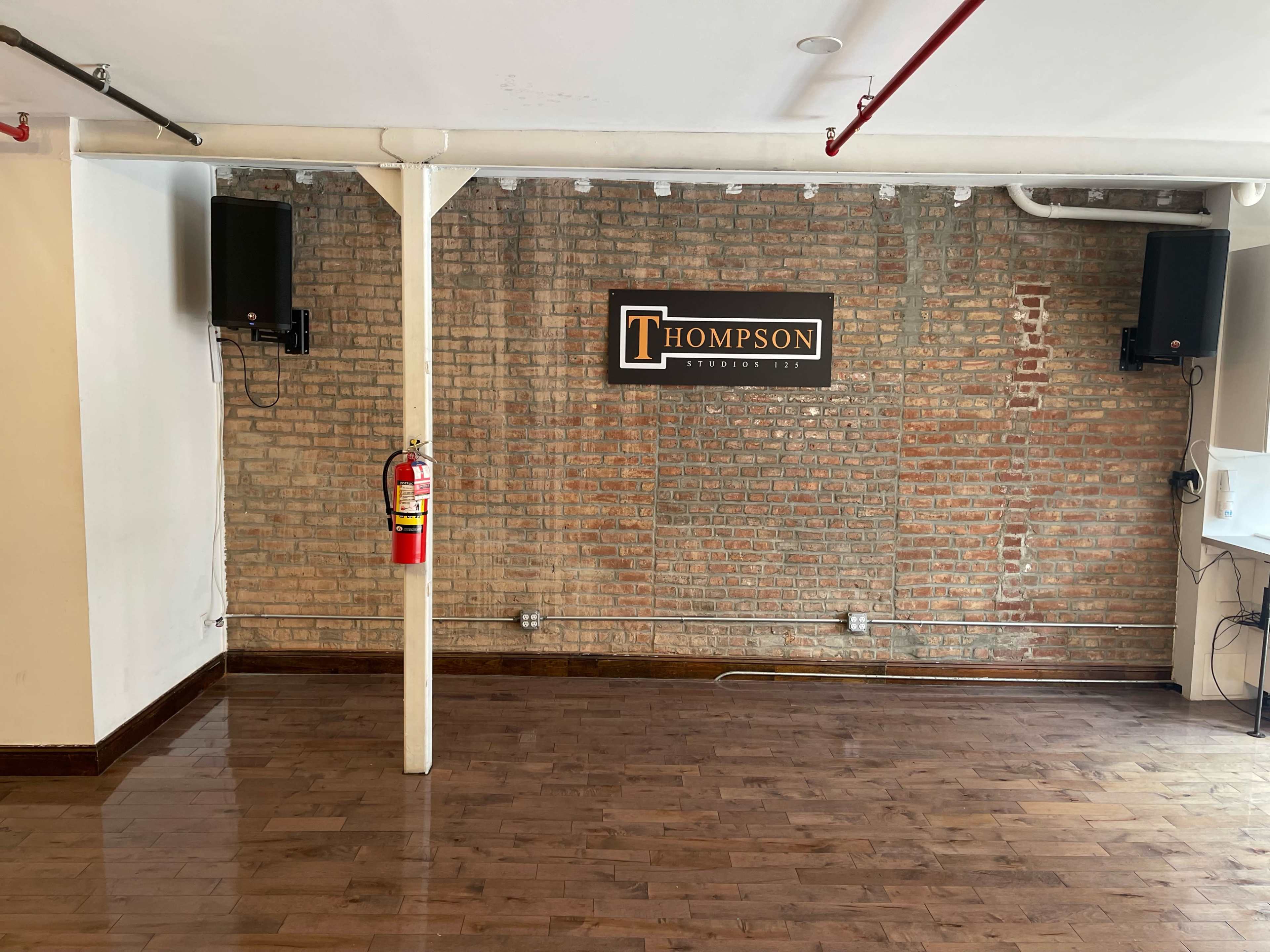 An empty room with a wooden floor, a brick wall featuring a sign that reads "THOMPSON," and audio speakers mounted on either side.