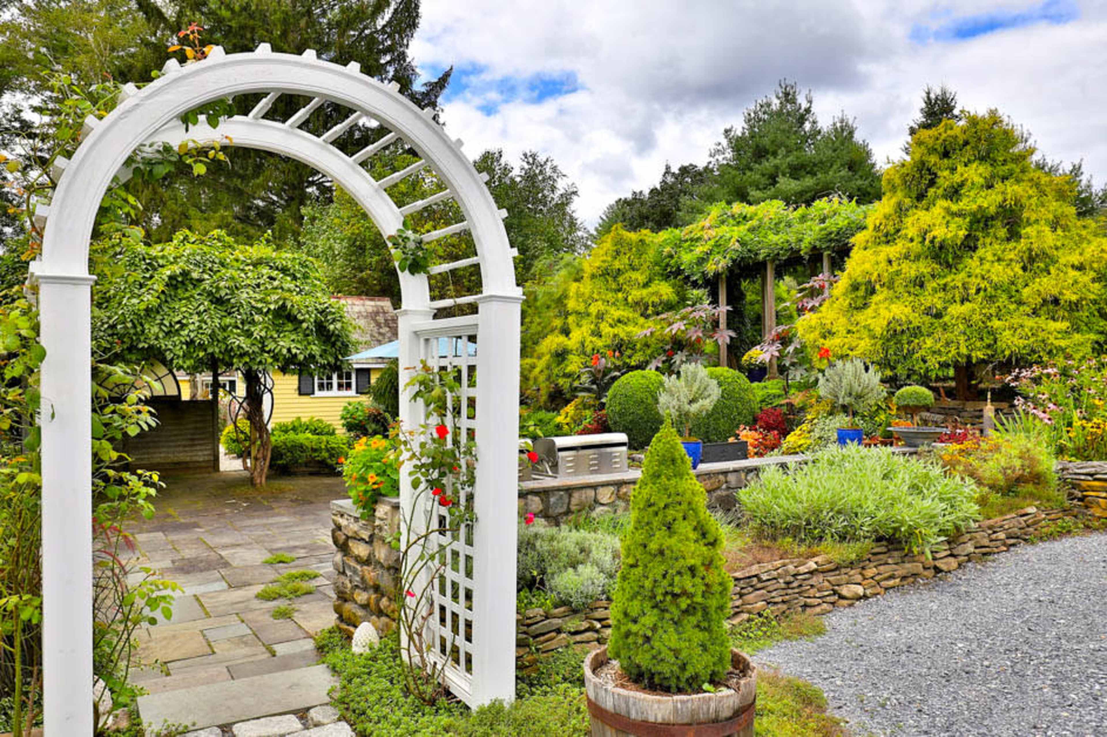 A white archway leads into a landscaped garden with various plants, shrubs, and stone pathways.