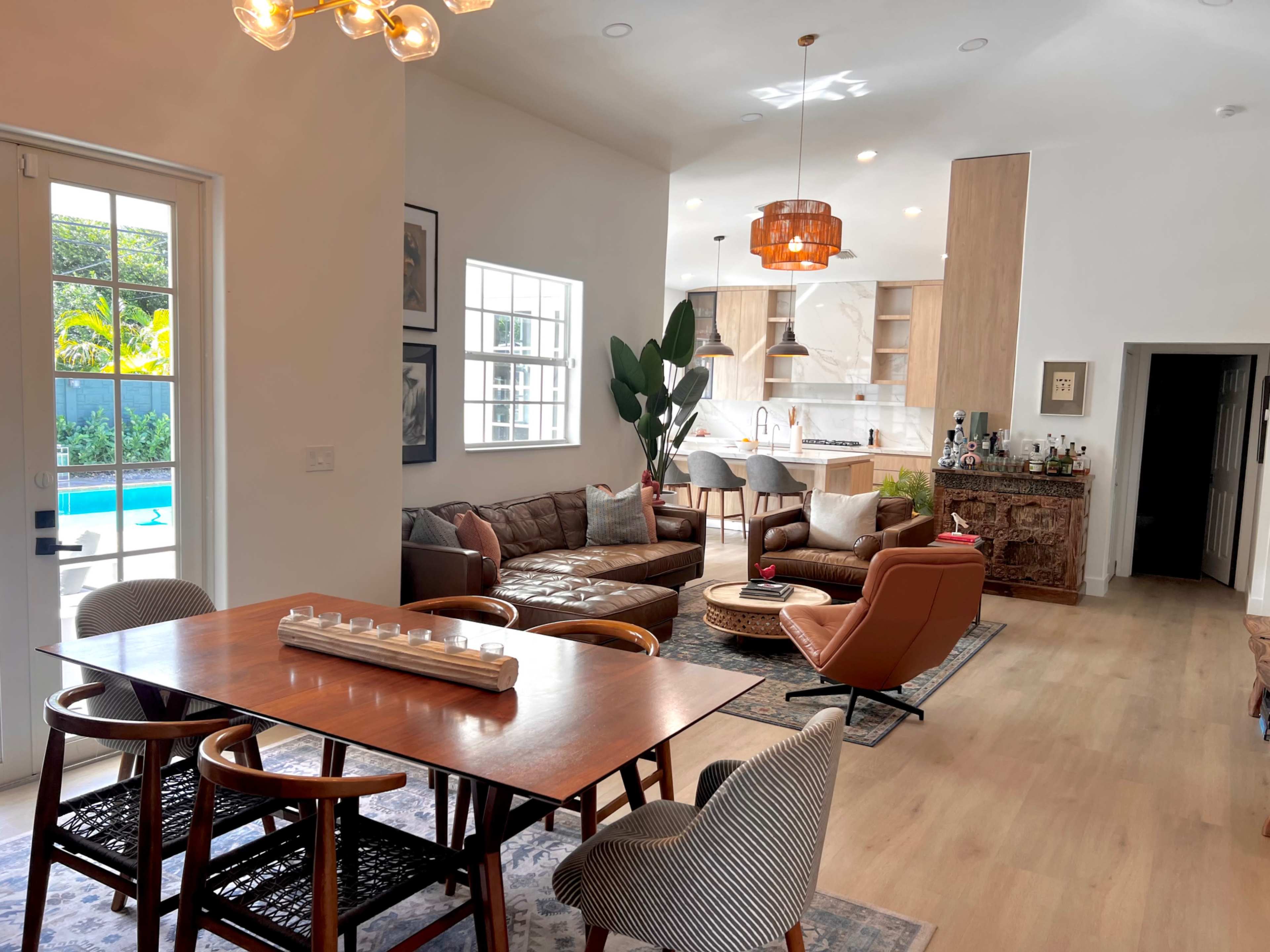 A modern living and dining area with a pool view, featuring a wooden dining table, a brown leather sofa, and a contemporary kitchen in the background.