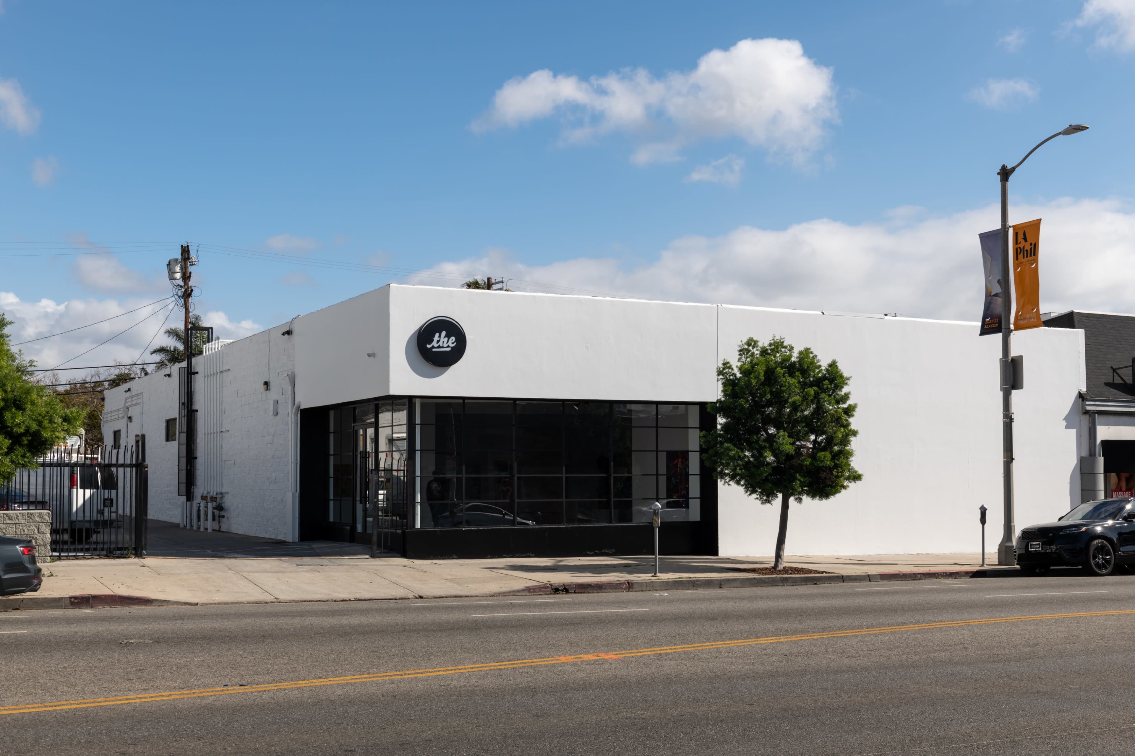 A modern white building with large windows and a circular logo, situated on a street next to a parked car.