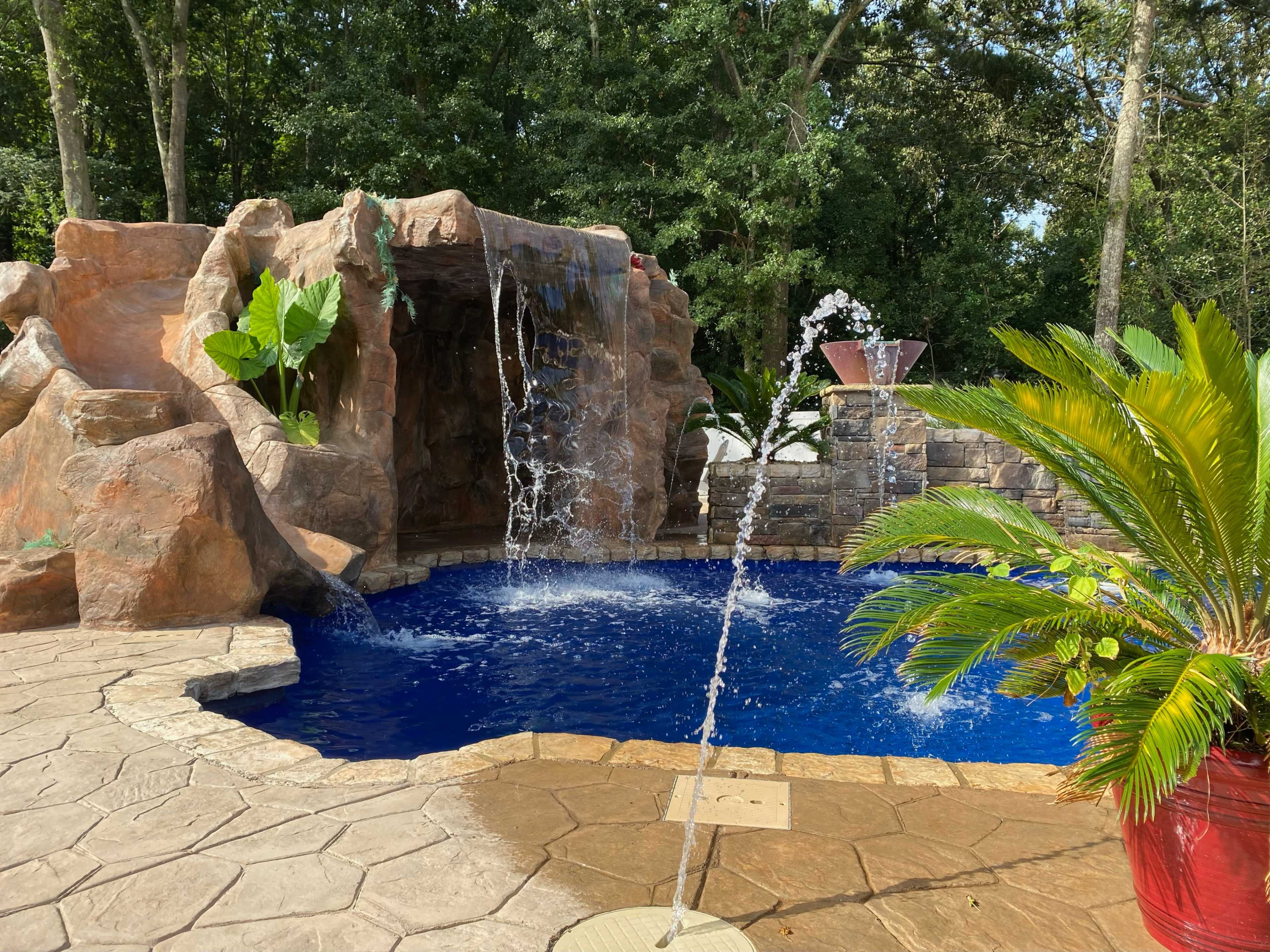 A landscaped pool area with a rock formation, waterfalls, and tropical plants surrounding a blue water surface.