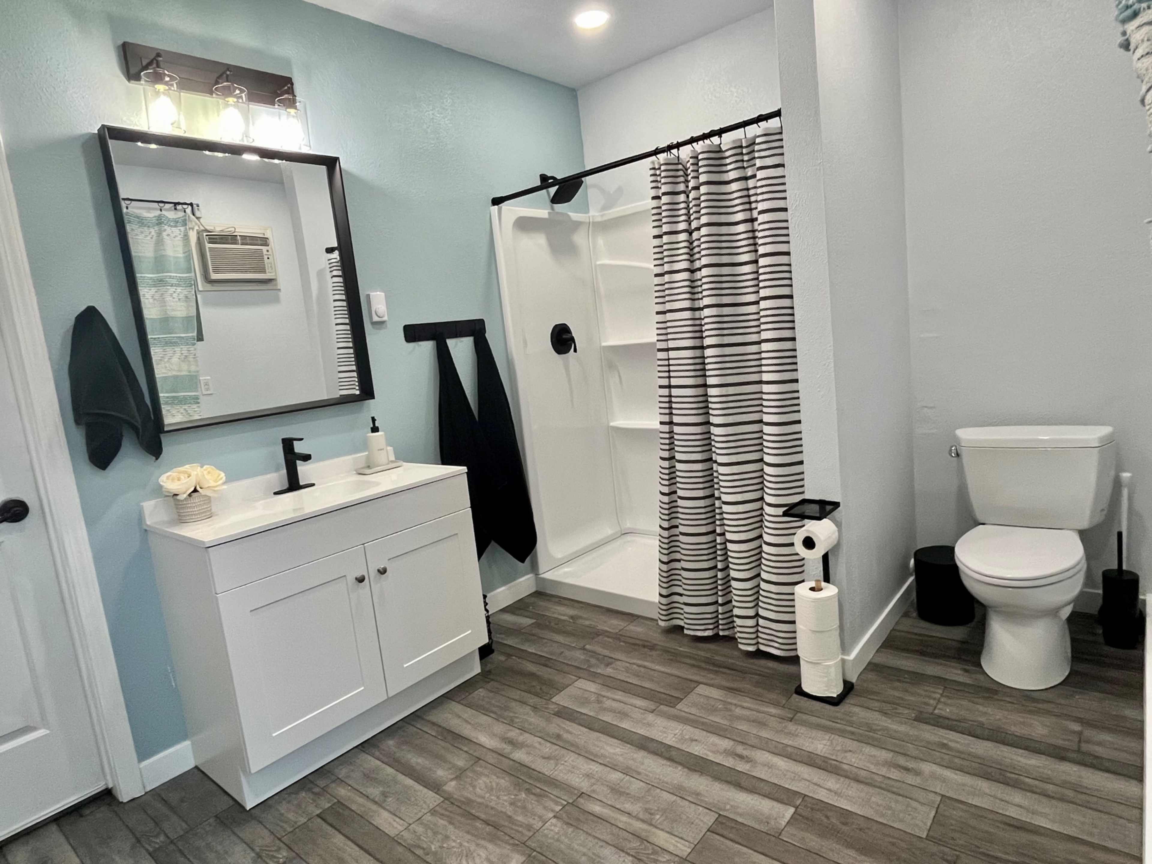 The bathroom features a shower with a striped curtain, a white vanity with a mirror, and a toilet, all set against light blue walls and wood-like flooring.