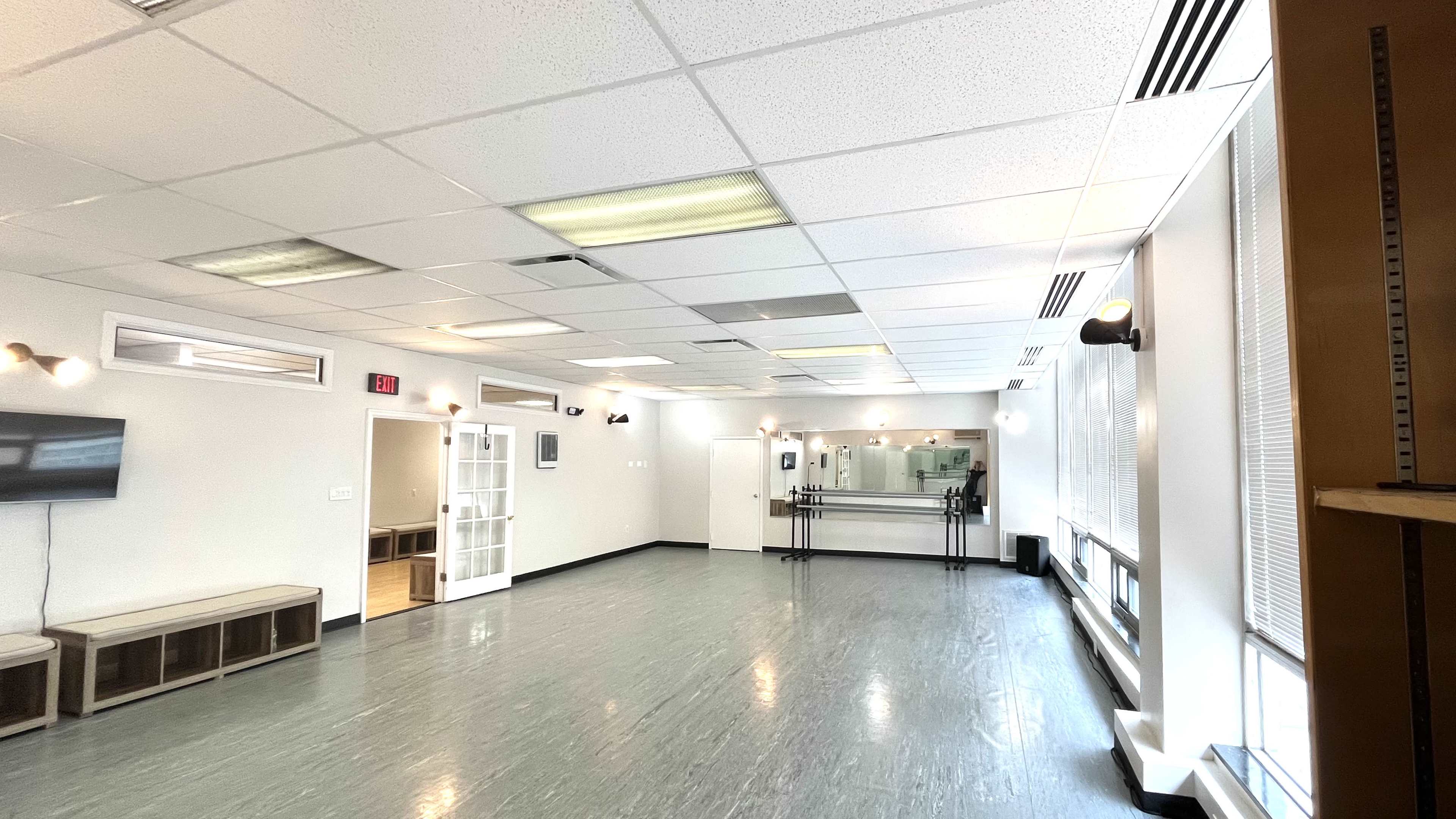 A spacious, empty room with gray flooring, white walls, and a large mirror at the far end, along with a bench and a television mounted on the wall.