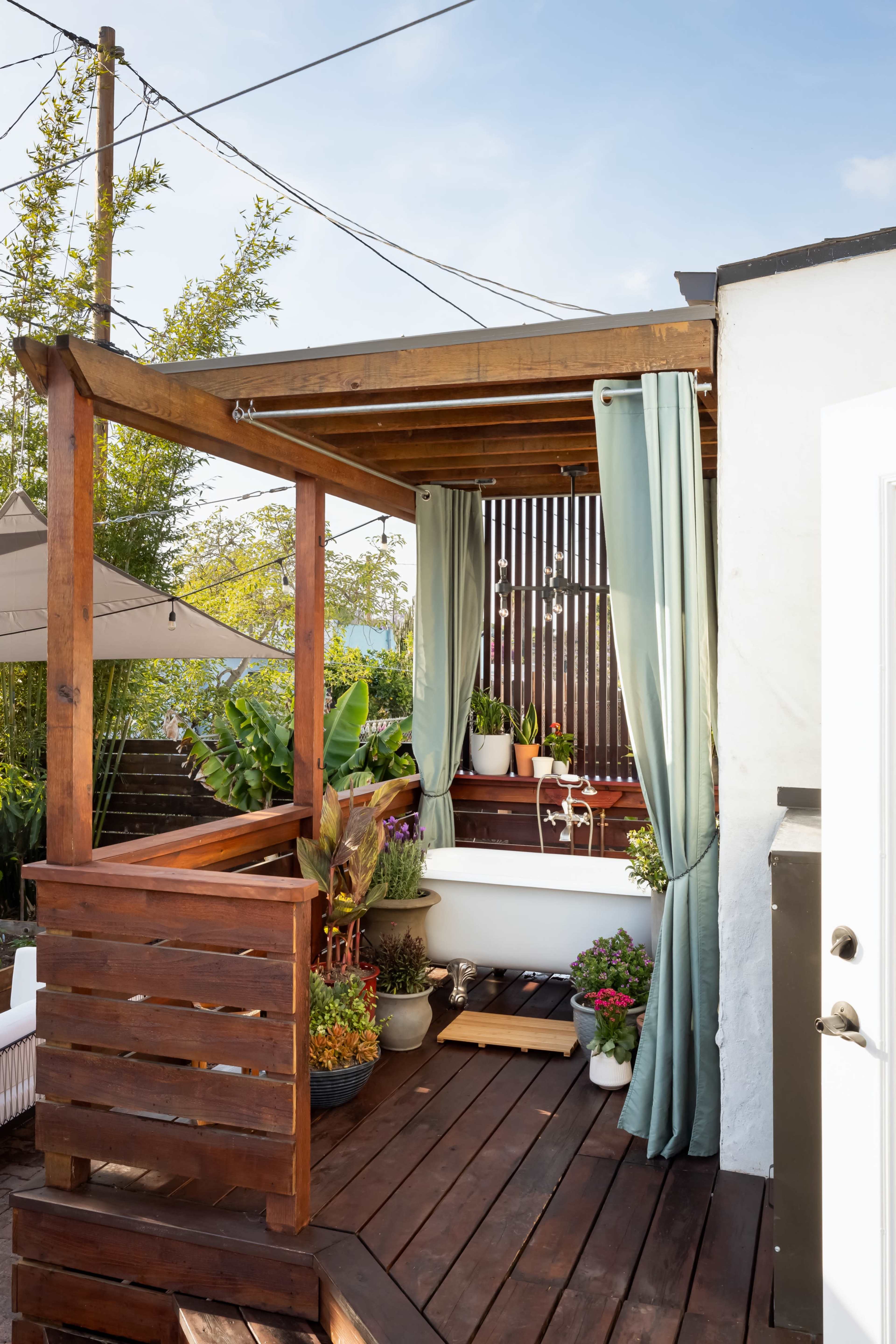 A wooden deck features a covered seating area with potted plants and curtains, surrounded by greenery and power lines.
