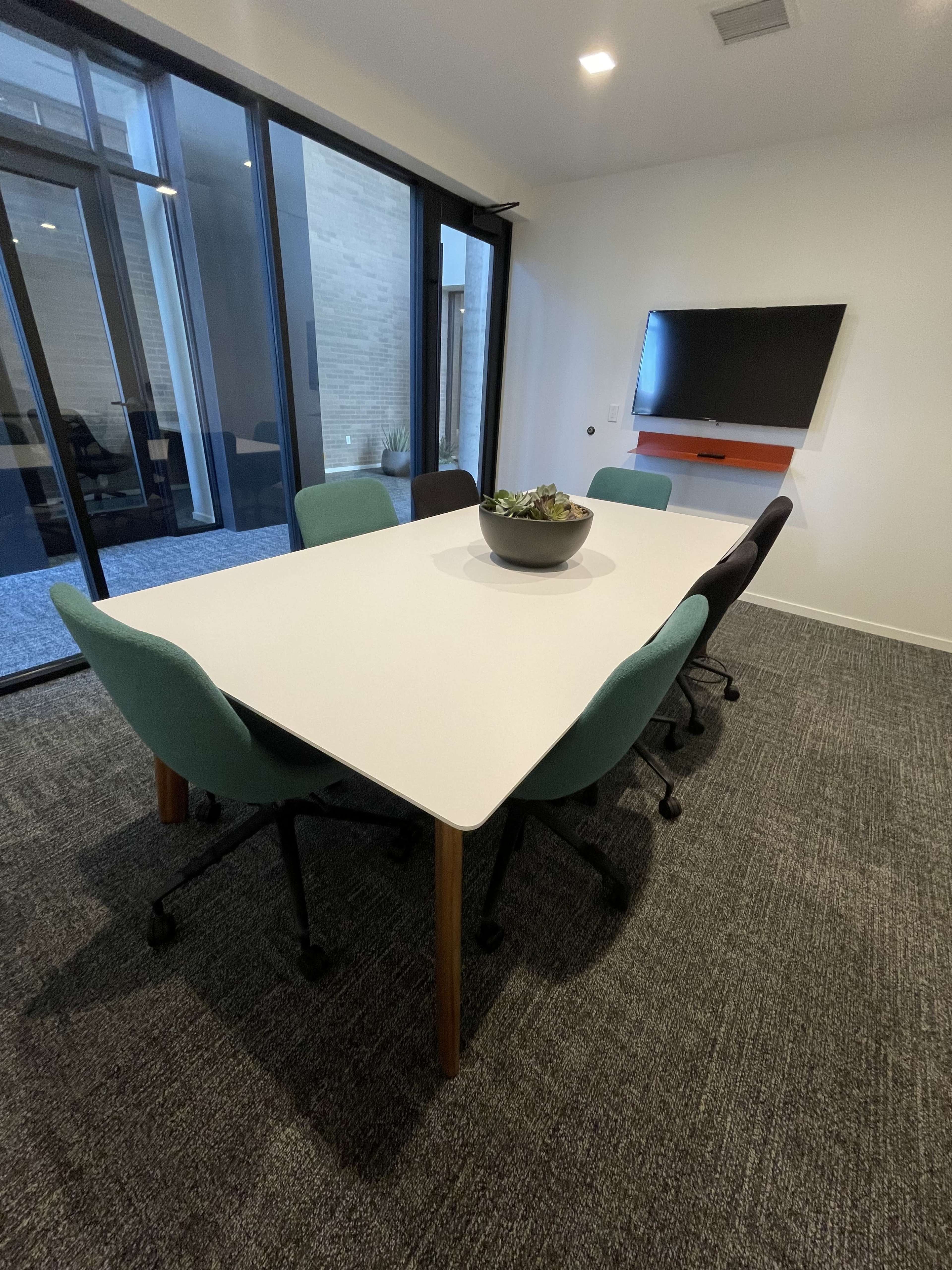 A conference room features a white table surrounded by chairs in various colors and a potted plant in the center, with large windows and a wall-mounted screen.