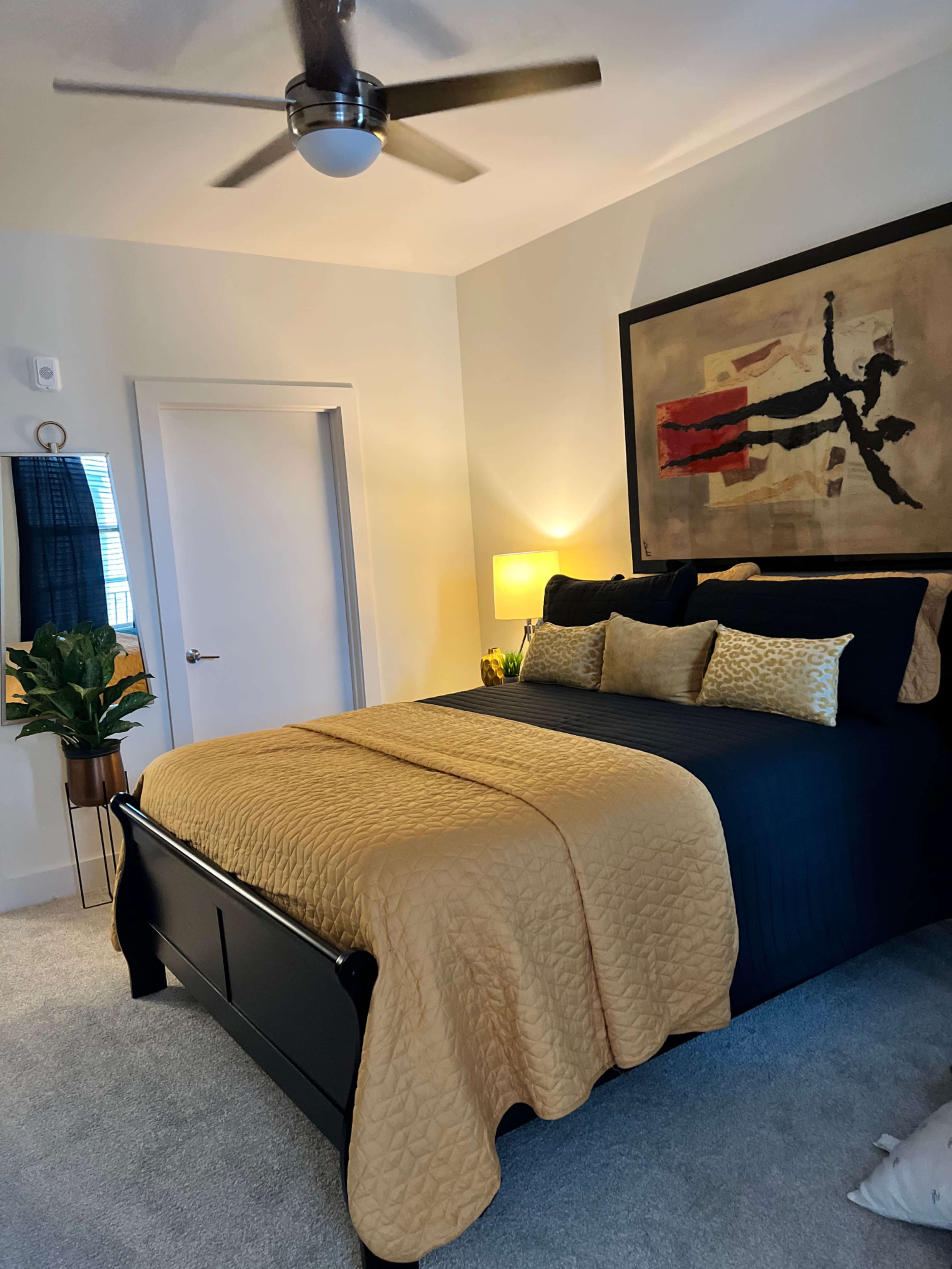 A well-decorated bedroom featuring a large bed with a black and gold color scheme, a framed artwork above the bed, and a potted plant in the corner.
